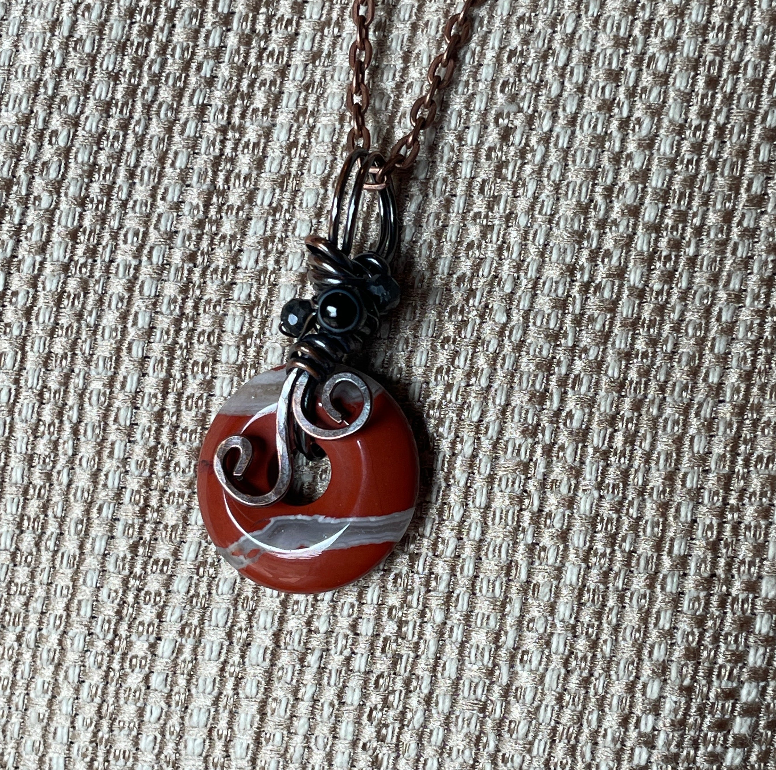 A handmade pendant necklace featuring a round stone disc charm with red, white, and gray swirls, wrapped in dark metal and hanging from a brown chain on a textured beige background.