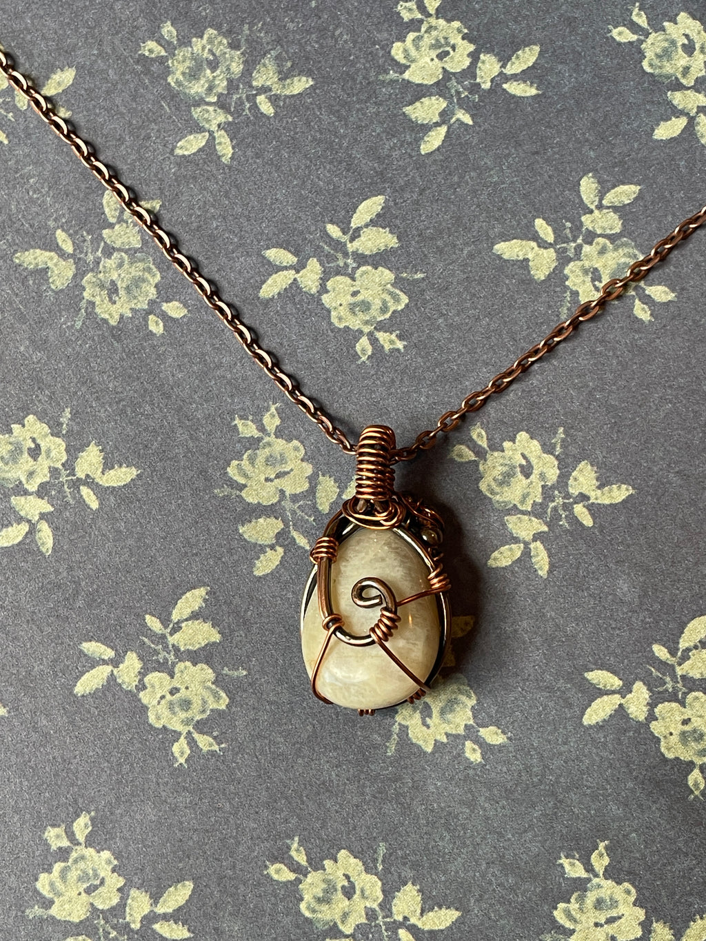 A spiral ammolite pendant wrapped in copper wire, hanging from a copper chain against a gray floral fabric background.