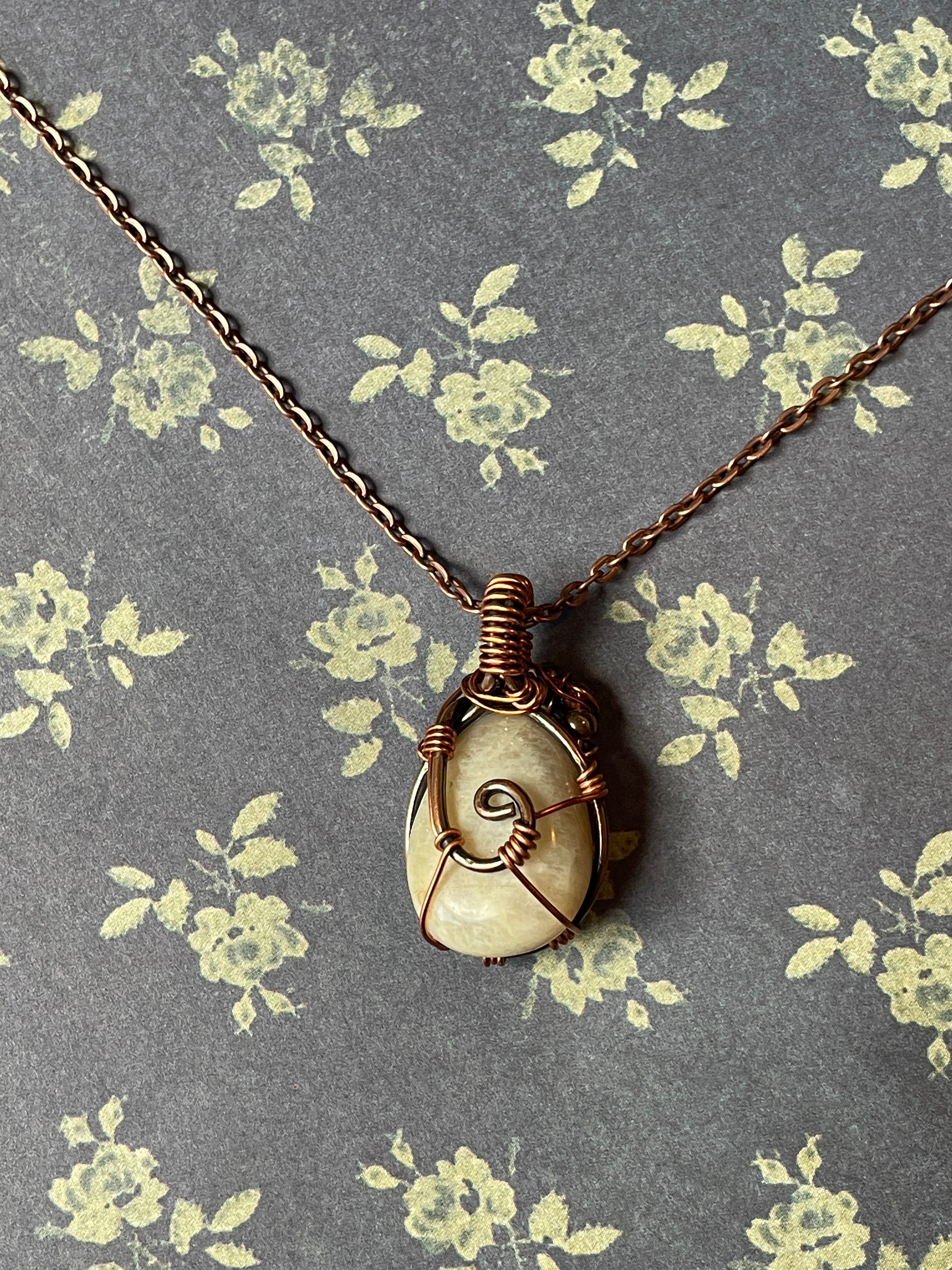 A spiral ammolite pendant wrapped in copper wire, hanging from a copper chain against a gray floral fabric background.