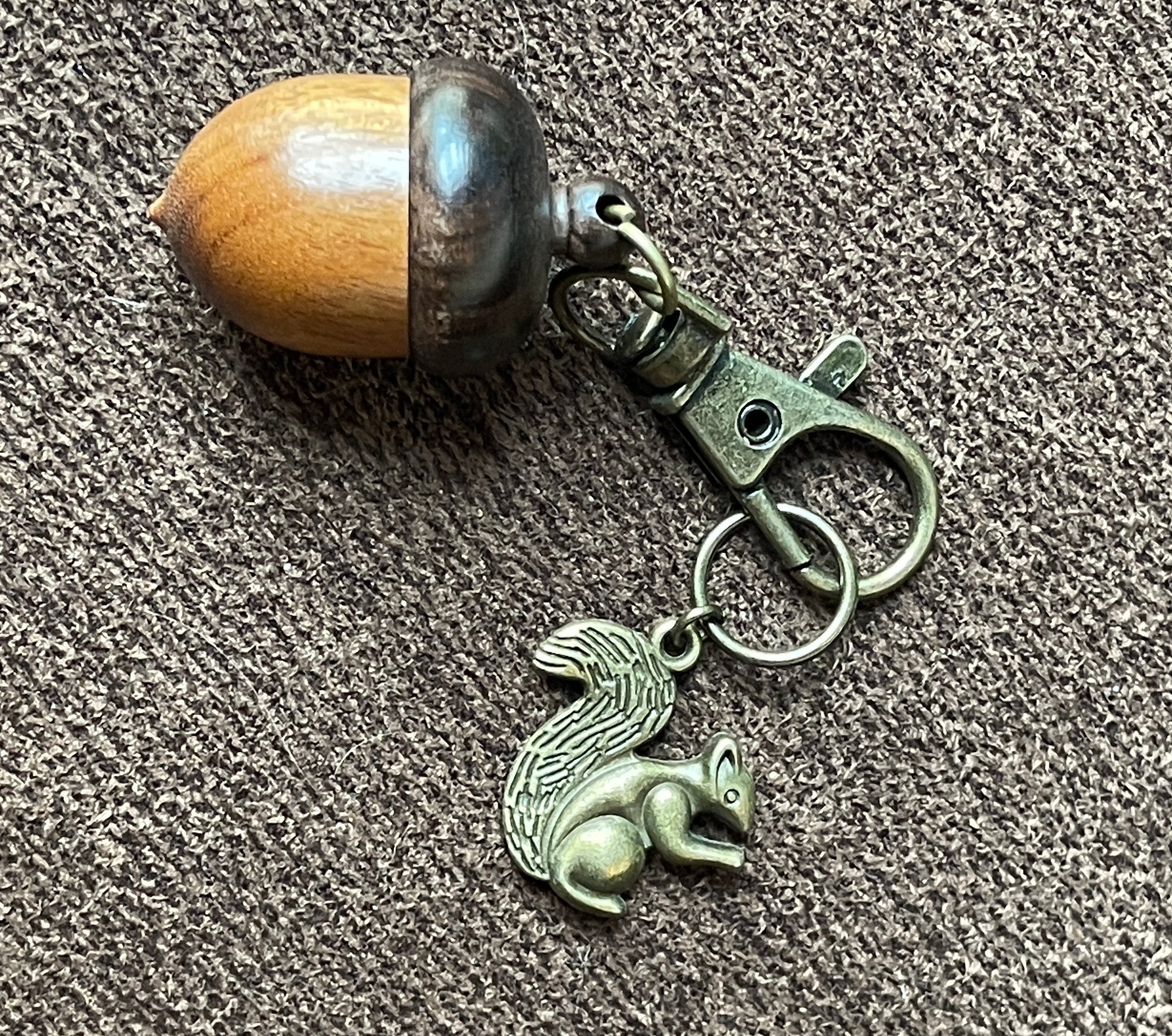 A keychain featuring a small metallic mermaid charm and a wooden acorn bobble on a split ring and clasp, shown on a textured gray carpet.