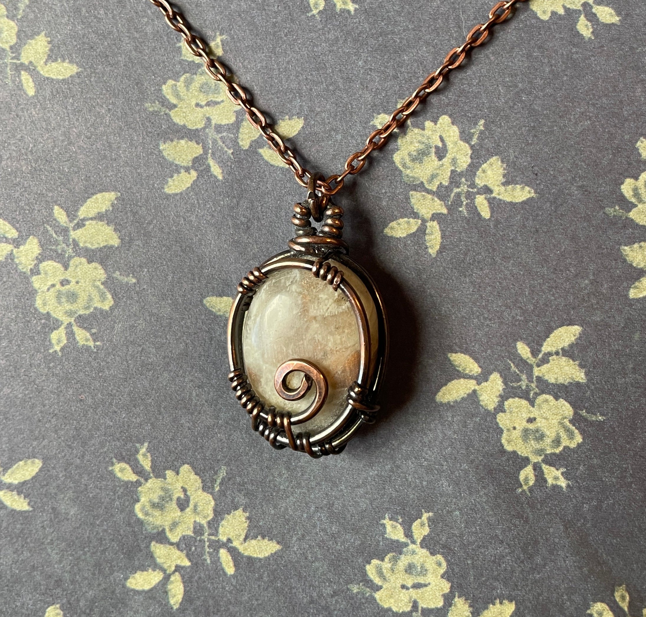 A handmade spiral shell pendant necklace with a rustic bronze wire-wrapped setting and a dark bronze chain, displayed on a gray background with a yellow floral pattern.