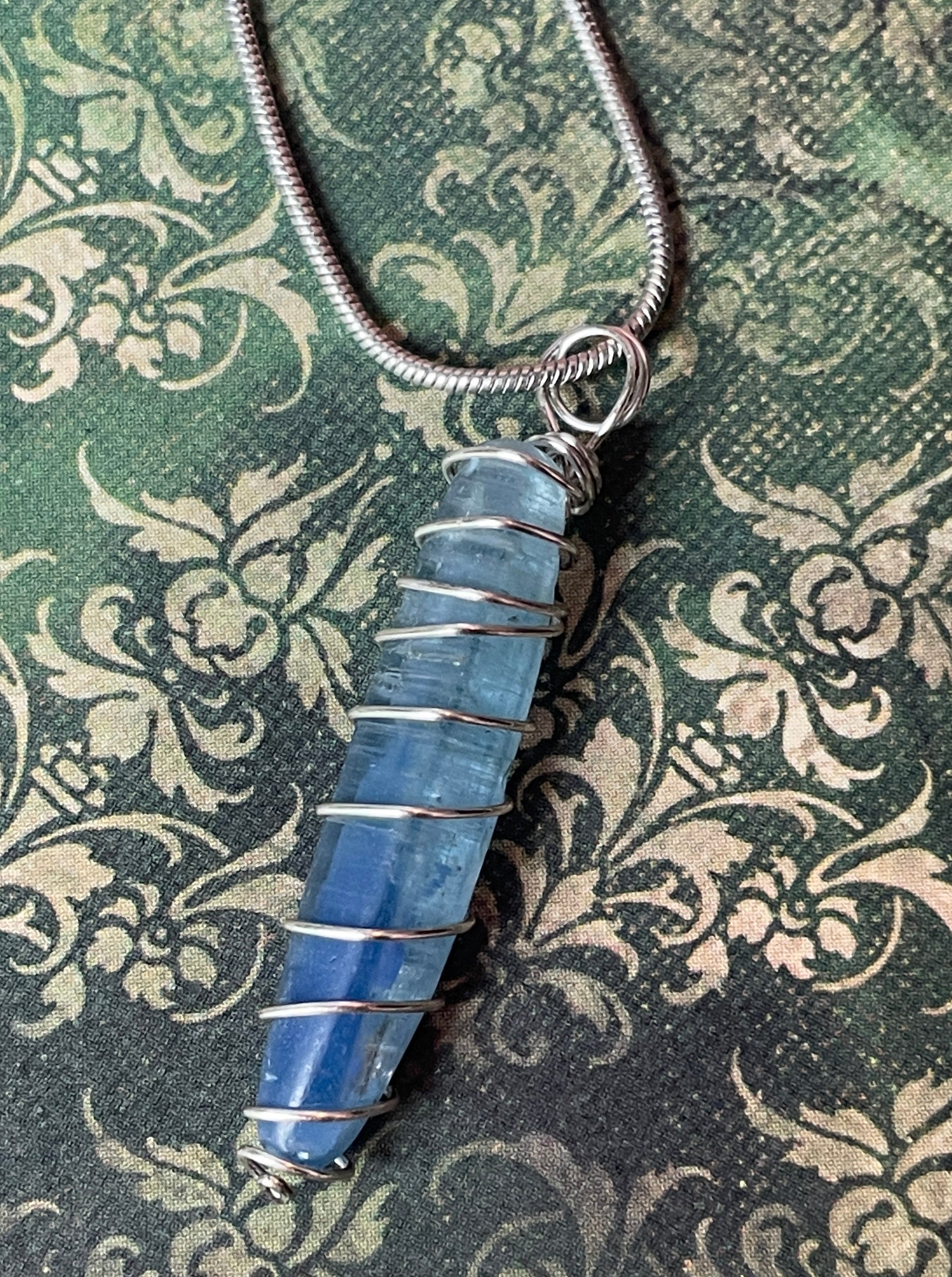 A blue stone pendant wrapped in silver wire, hanging from a silver snake chain, displayed on a dark green damask patterned fabric background.