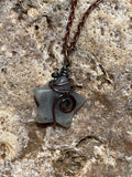 A handmade star-shaped pendant necklace made from a gray stone wrapped in dark copper wire, resting on a textured beige rock surface.
