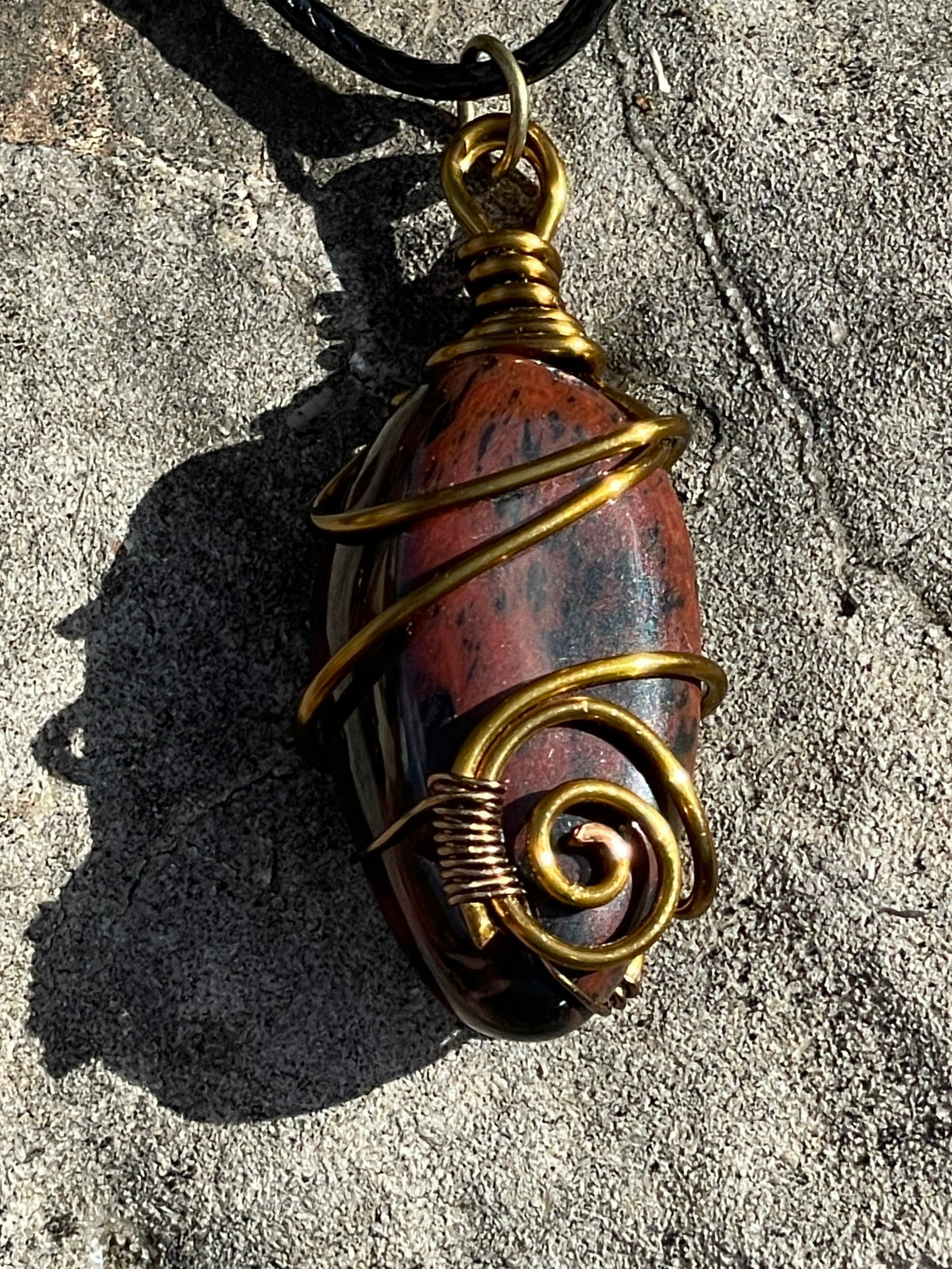 A handmade wire-wrapped pendant necklace featuring a polished reddish-brown stone with dark inclusions, wrapped in gold-toned metal and hanging from a black cord on a textured gray stone surface.