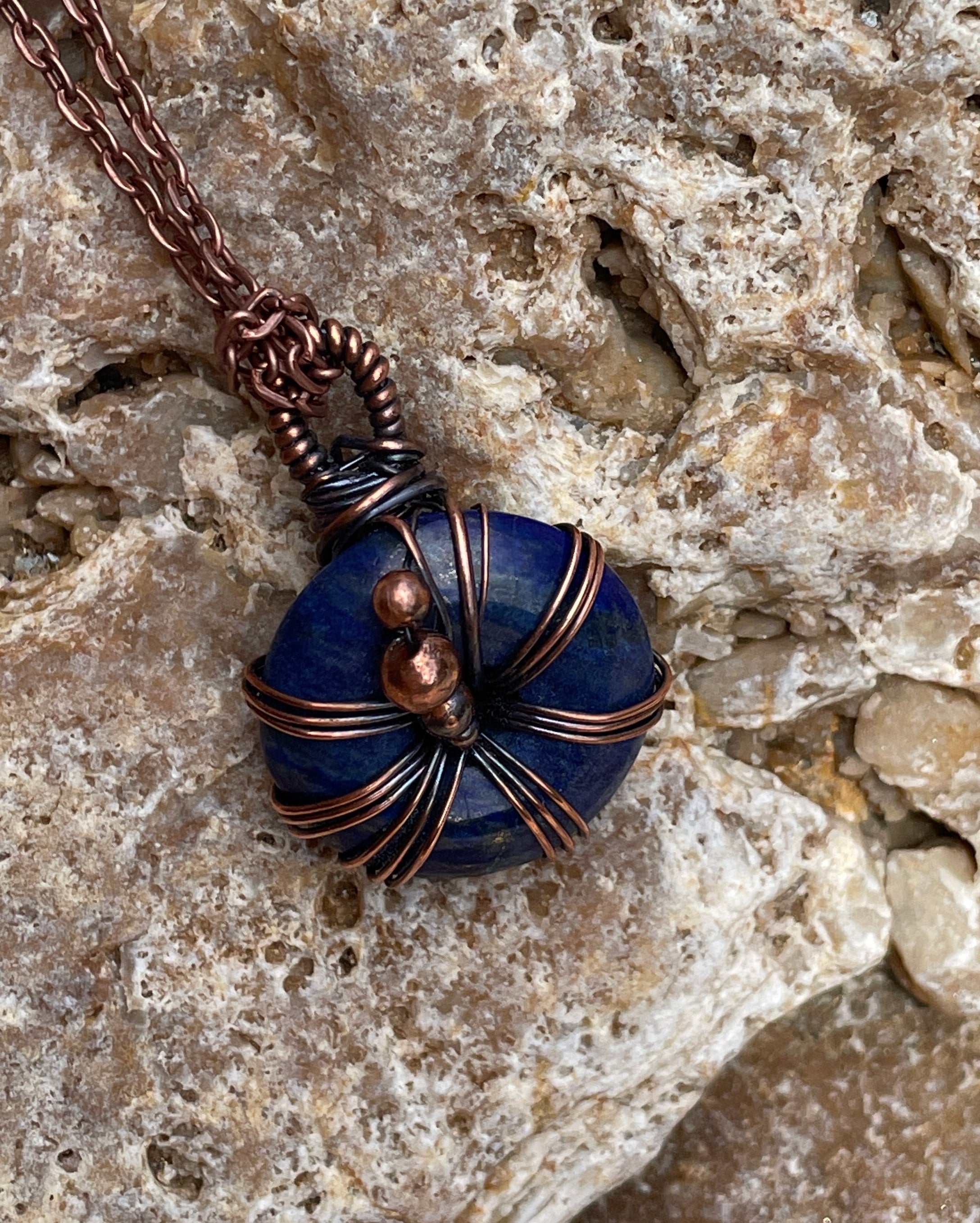 A handmade pendant necklace featuring a deep blue stone bead wrapped in copper wire, resting on a textured light gray rock surface.