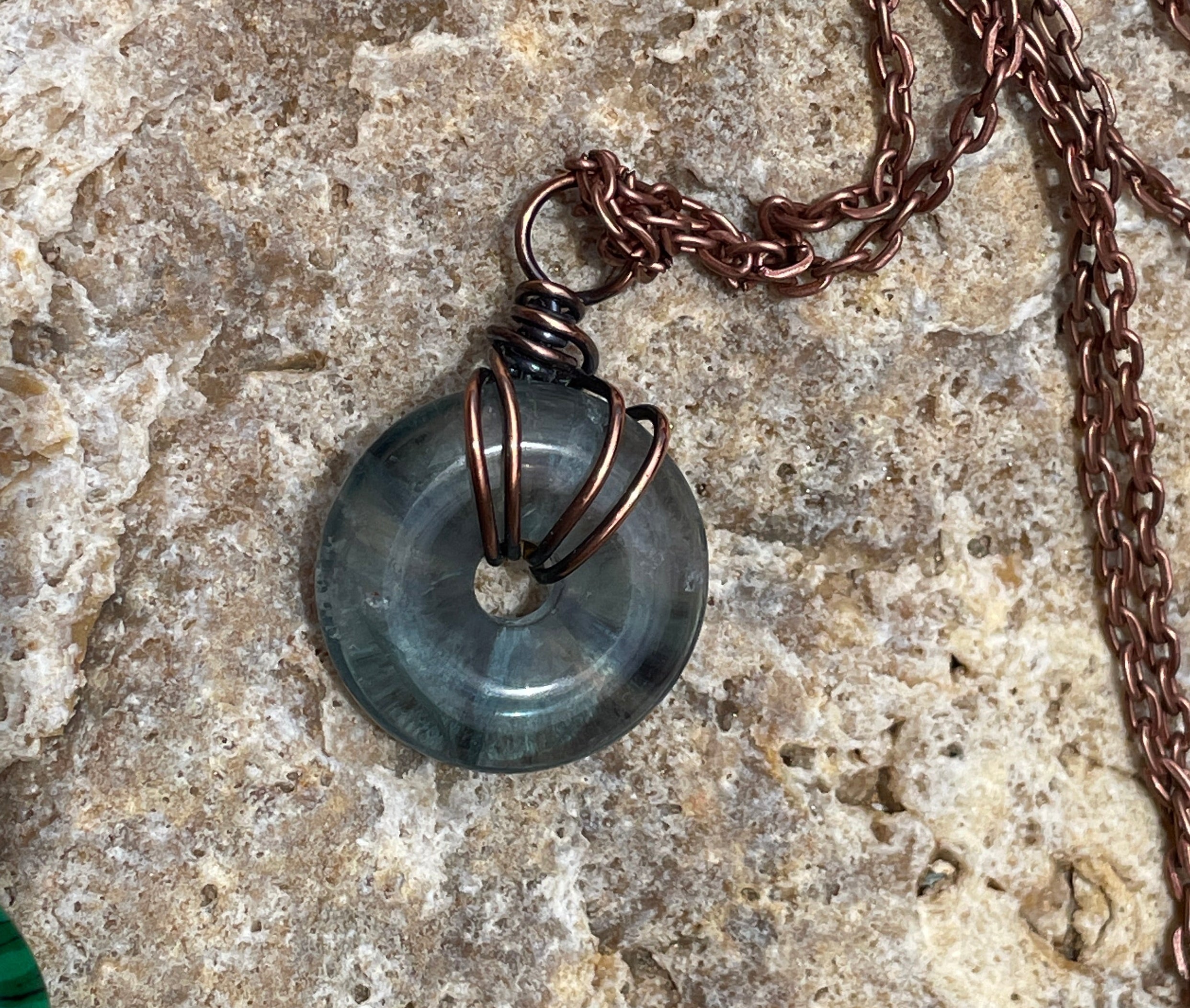 A close-up of a handmade necklace featuring a round, dark gray stone pendant wrapped in copper wire, resting on a textured beige stone surface.