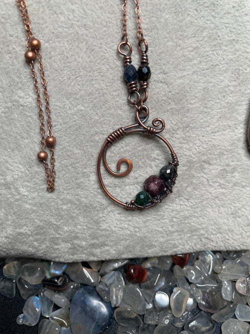 A handmade copper wire pendant necklace with a spiral design and small multicolor gemstone beads, displayed on a textured gray fabric next to polished stones.