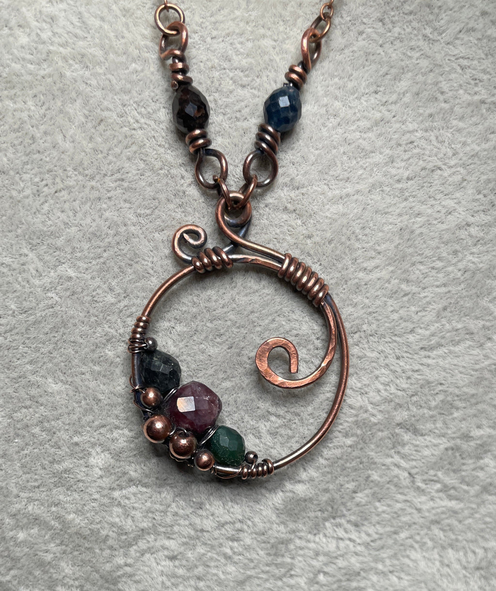 A close-up of a handmade copper wire pendant necklace with spiral details and small faceted beads in purple, green, and dark blue, displayed on a textured gray fabric next to a bed of polished stones.