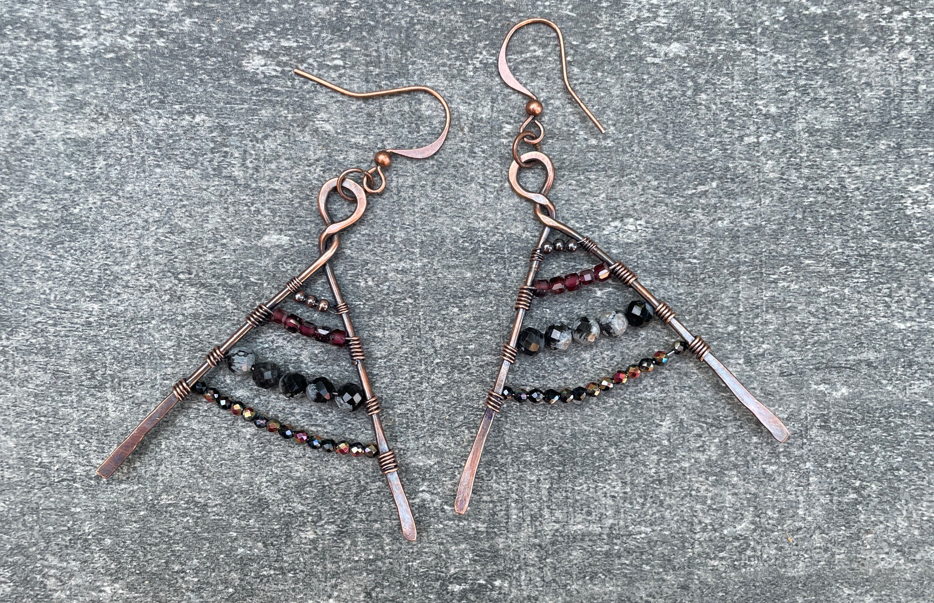 A pair of handmade dangle earrings with copper-toned metal hooks and triangular wire frames, decorated with small dark beads, laid flat on a textured gray surface.