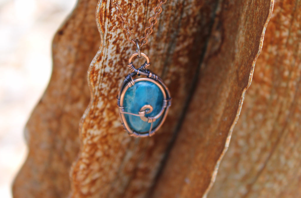 A close-up shot of a handmade pendant featuring a round turquoise-blue stone wrapped in textured copper wire, hanging against a background of rusted brown leaves.