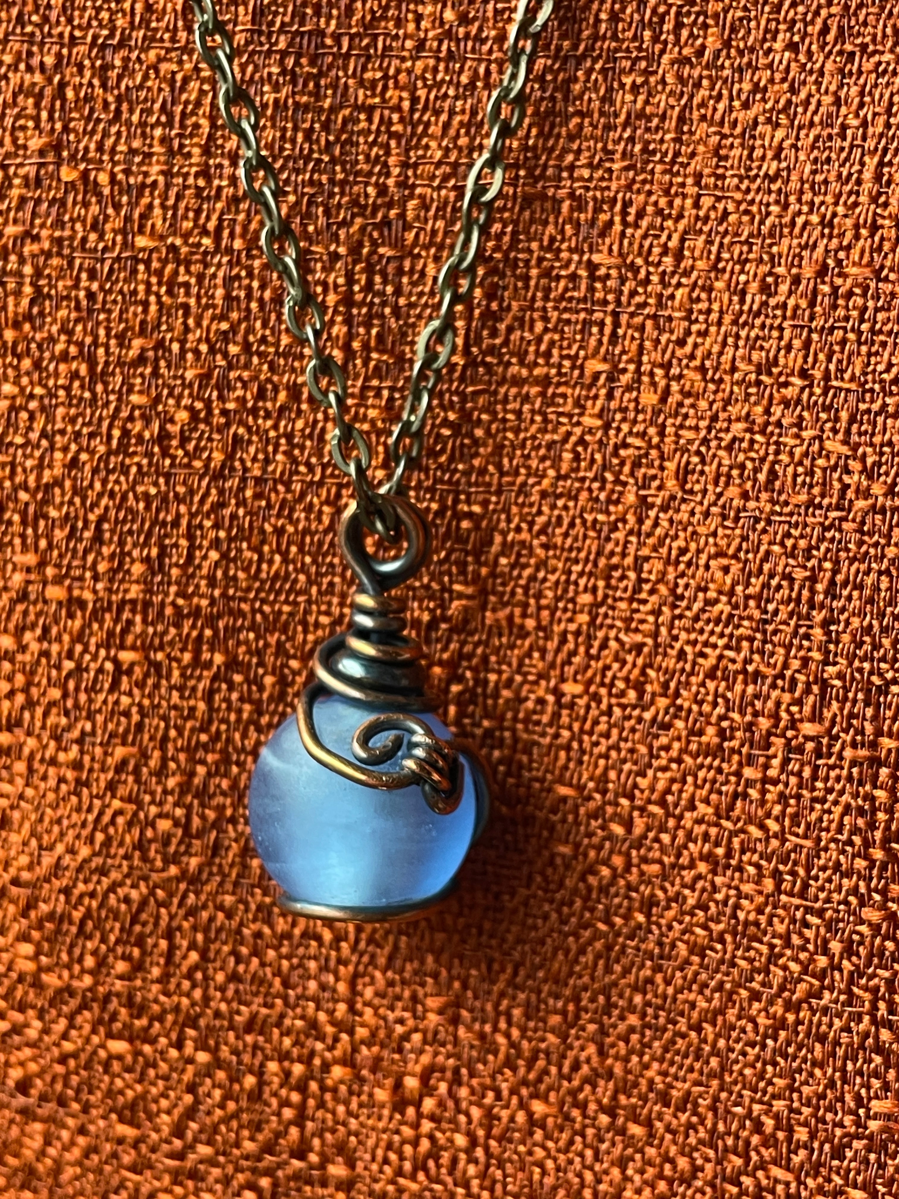 A close-up of a necklace with a small light blue glass bottle pendant wrapped in dark metal, hanging against a textured orange background.