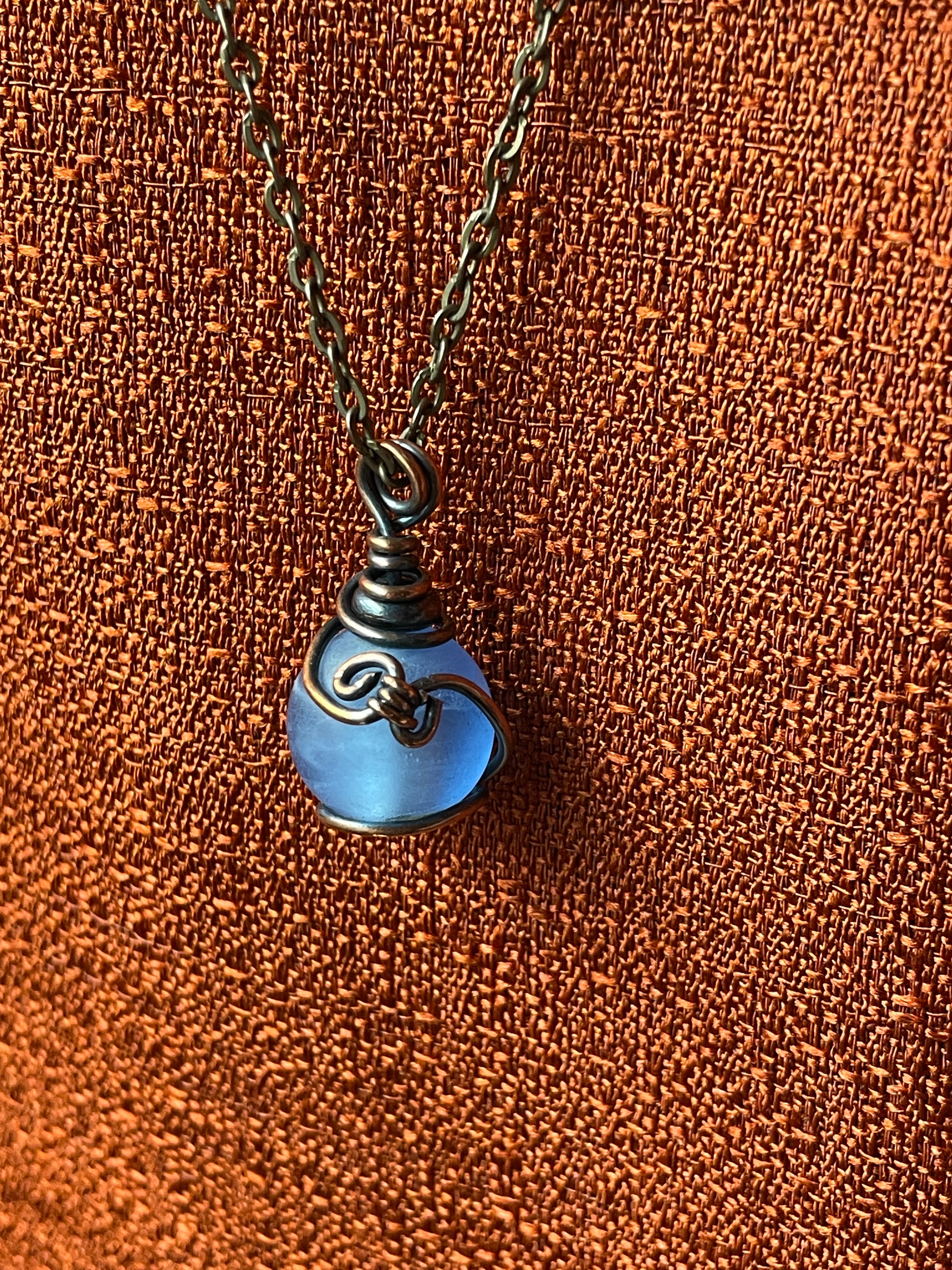 A close-up of a handmade pendant necklace featuring a light blue glass bead wrapped with dark bronze metal wire, hanging from a matching dark chain against a textured orange-brown background.