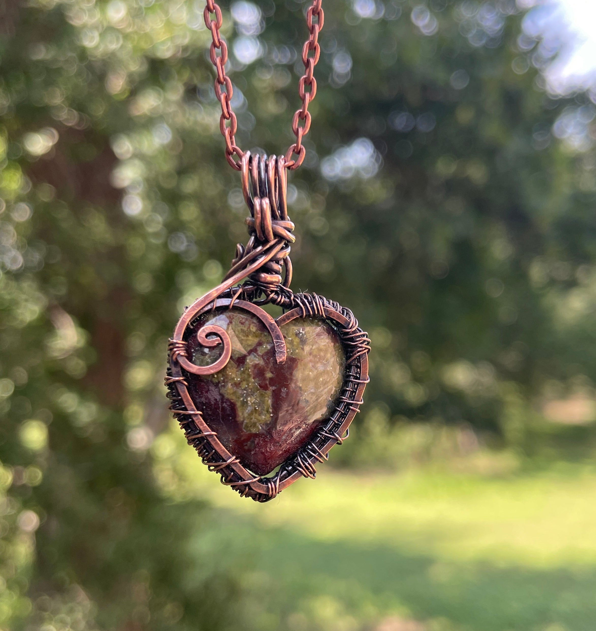 A heart-shaped pendant necklace made of copper wire wrapping around a natural stone, hanging against a blurred green outdoor background.