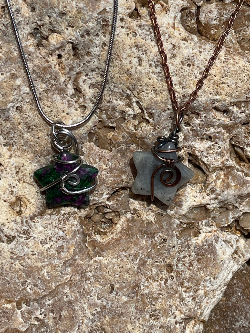 Two handmade wire-wrapped stone pendants on chains, displayed on a textured natural rock surface. One pendant is wrapped in silver wire with a green and purple stone, and the other is wrapped in copper wire with a gray stone.