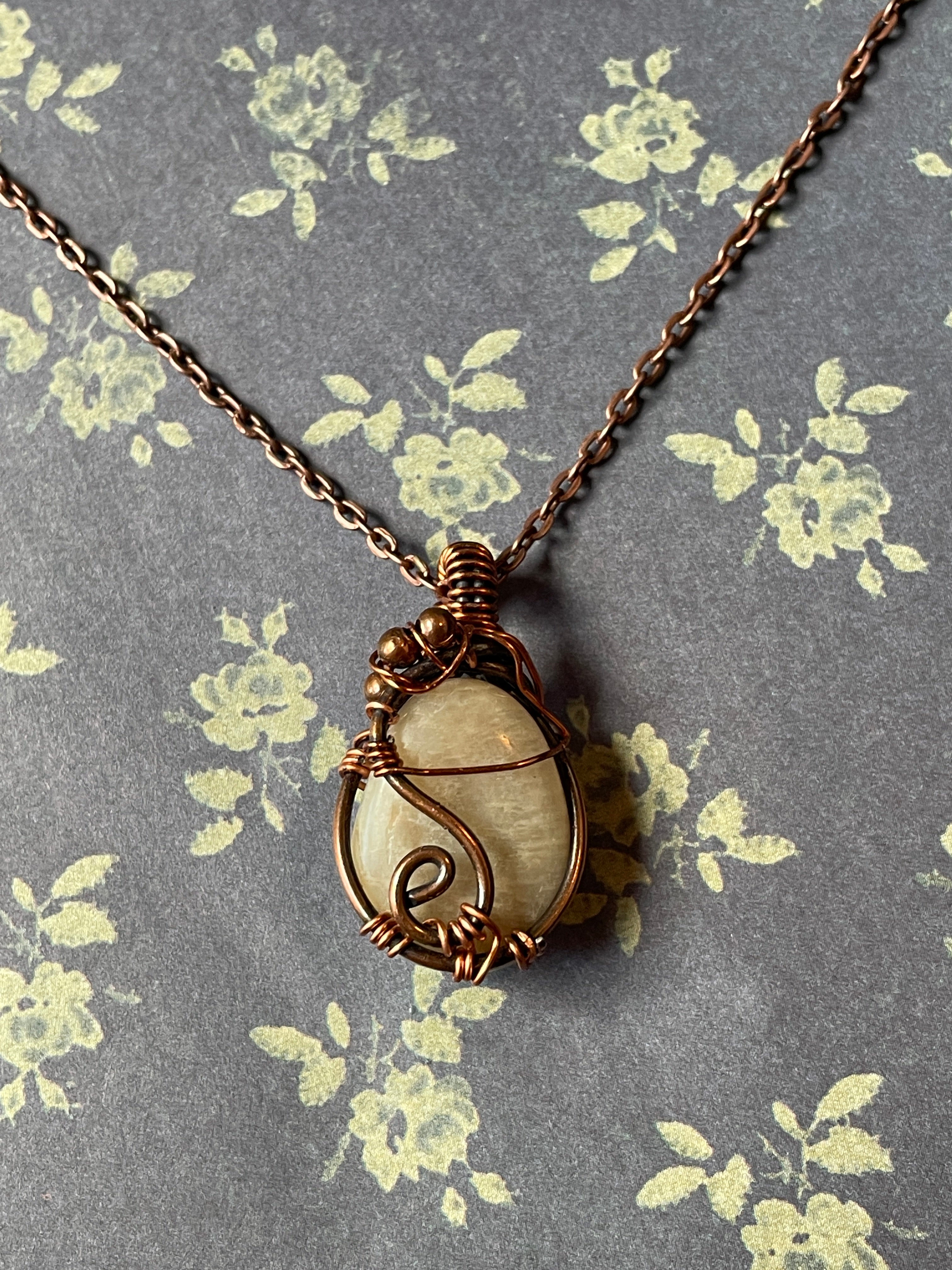 A handmade pendant necklace featuring a round cream-colored stone wrapped in copper wire, resting on a gray floral-patterned background.