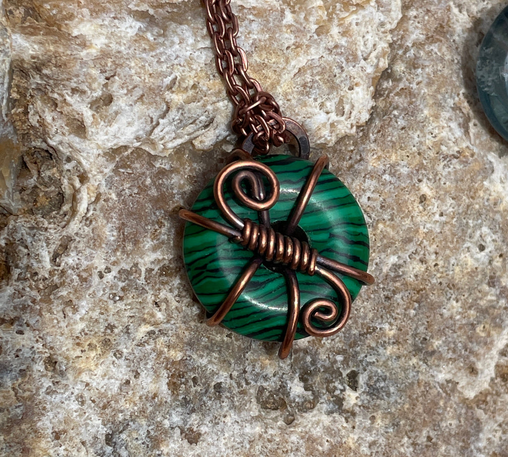 A close-up of a handmade pendant necklace featuring a green and black banded stone wrapped in copper wire, resting on a textured beige stone surface.