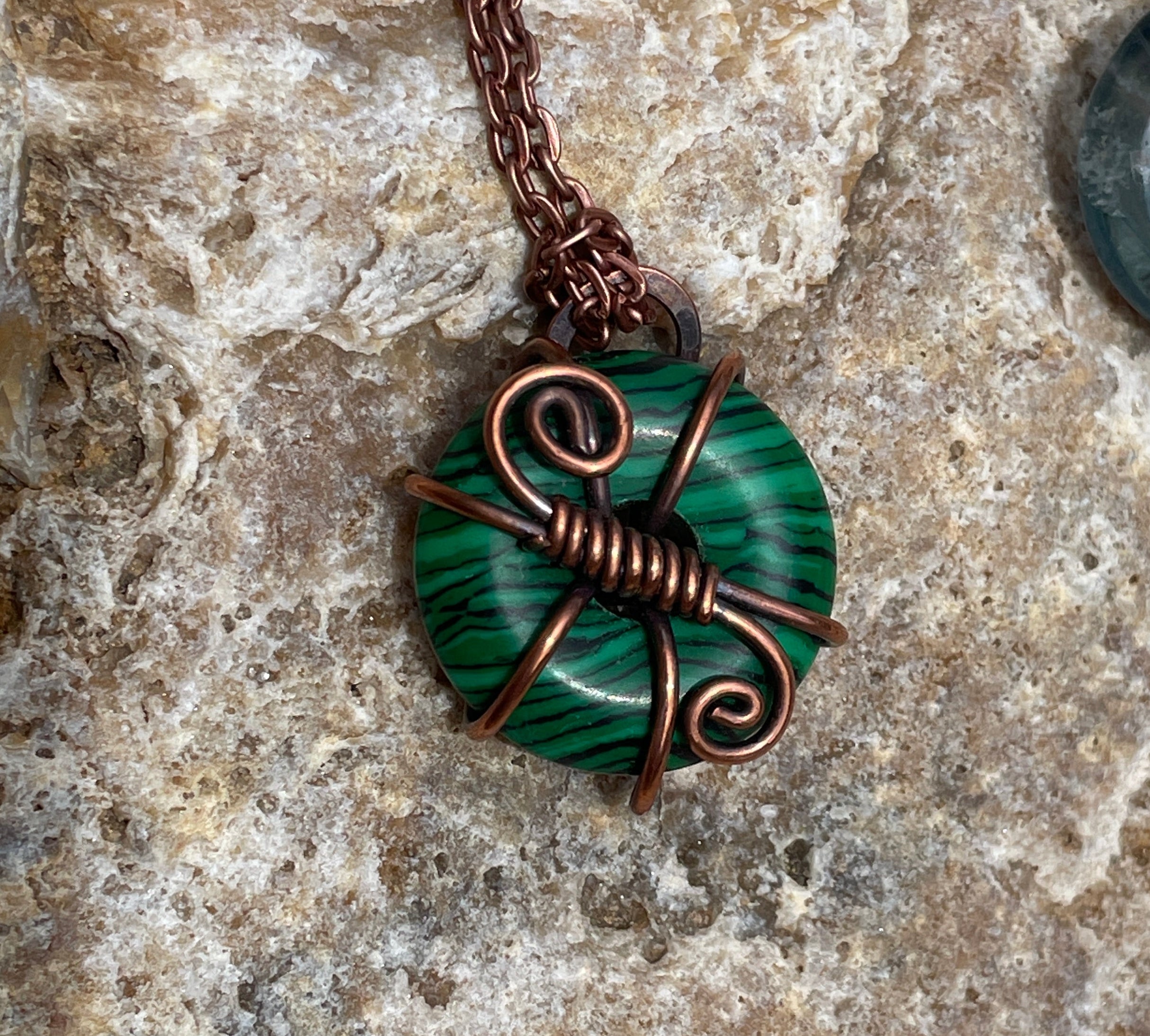 A close-up of a handmade pendant necklace featuring a green and black banded stone wrapped in copper wire, resting on a textured beige stone surface.