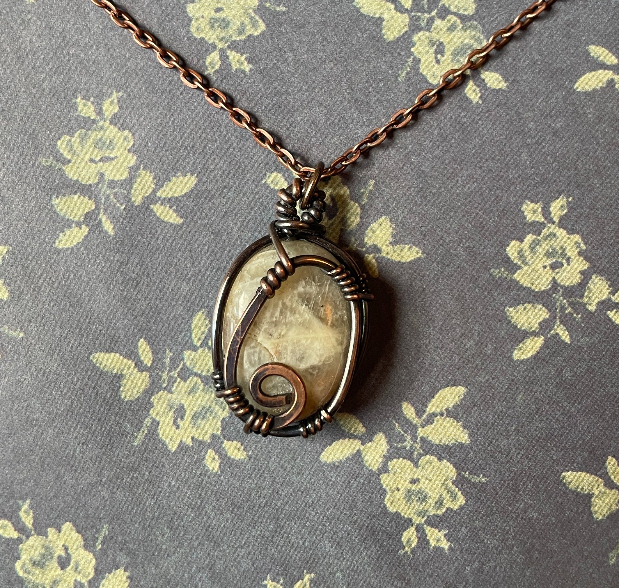 A handmade oval pendant necklace featuring a natural seashell wrapped in dark bronze and copper wire, hanging from a copper chain against a gray floral background.