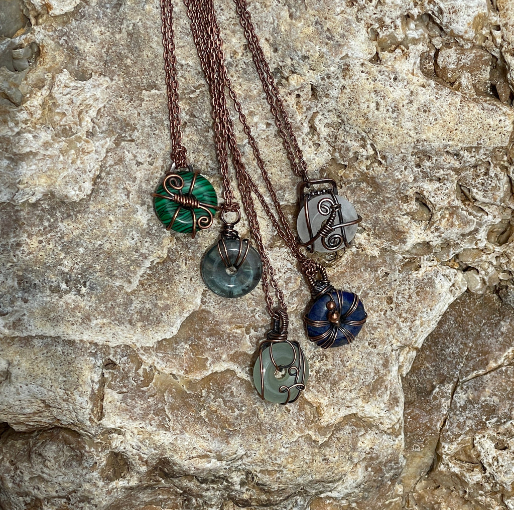 Five handmade necklaces with copper colored chains and various stone pendants in green, blue, and gray, arranged on a textured light gray rock.