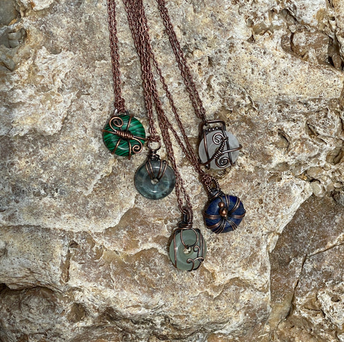 Five handmade necklaces with copper colored chains and various stone pendants in green, blue, and gray, arranged on a textured light gray rock.