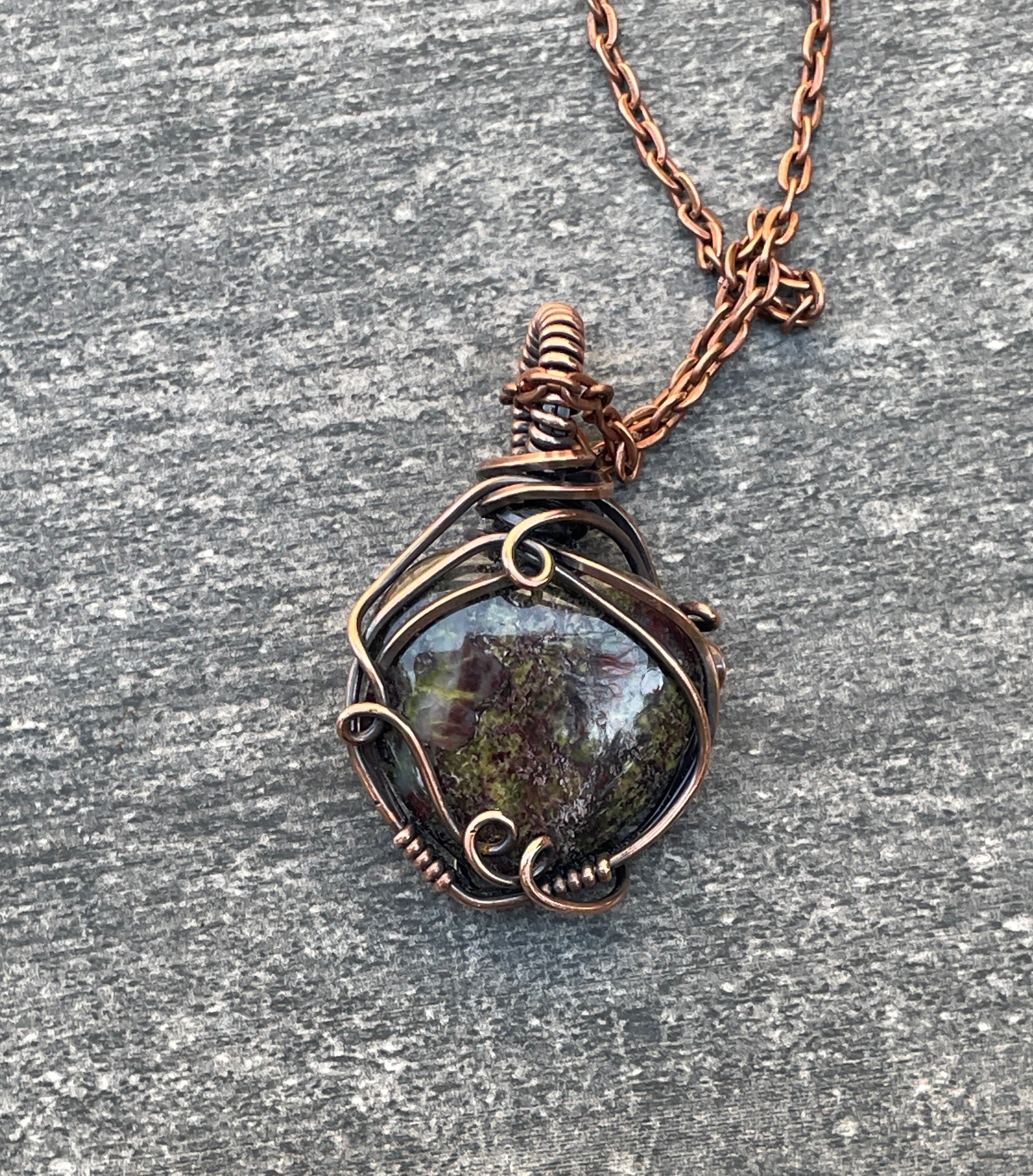 A handmade pendant necklace featuring a dark, polished stone wrapped in rustic copper wire, resting on a textured gray stone surface.