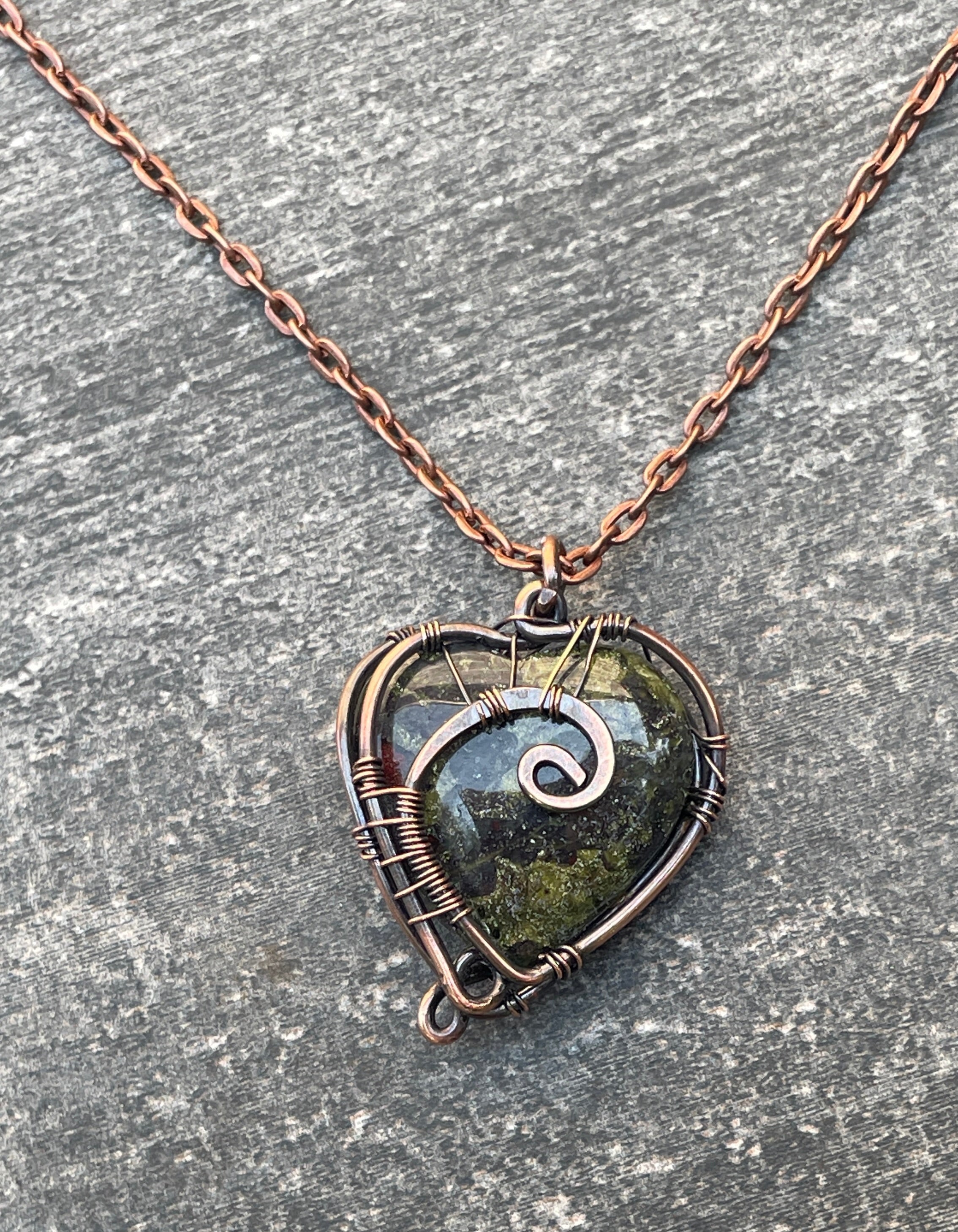 A heart-shaped wire-wrapped pendant necklace featuring a dark green stone with a silver spiral design, hanging from a copper-toned chain on a textured gray stone surface.