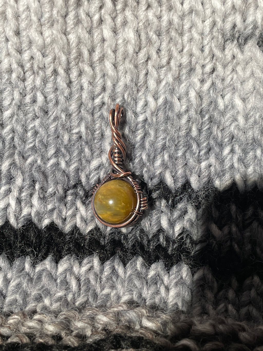 A small handmade wire-wrapped charm with a round olive-green stone and a twisted copper-toned bail, resting on a gray and black knit fabric background.