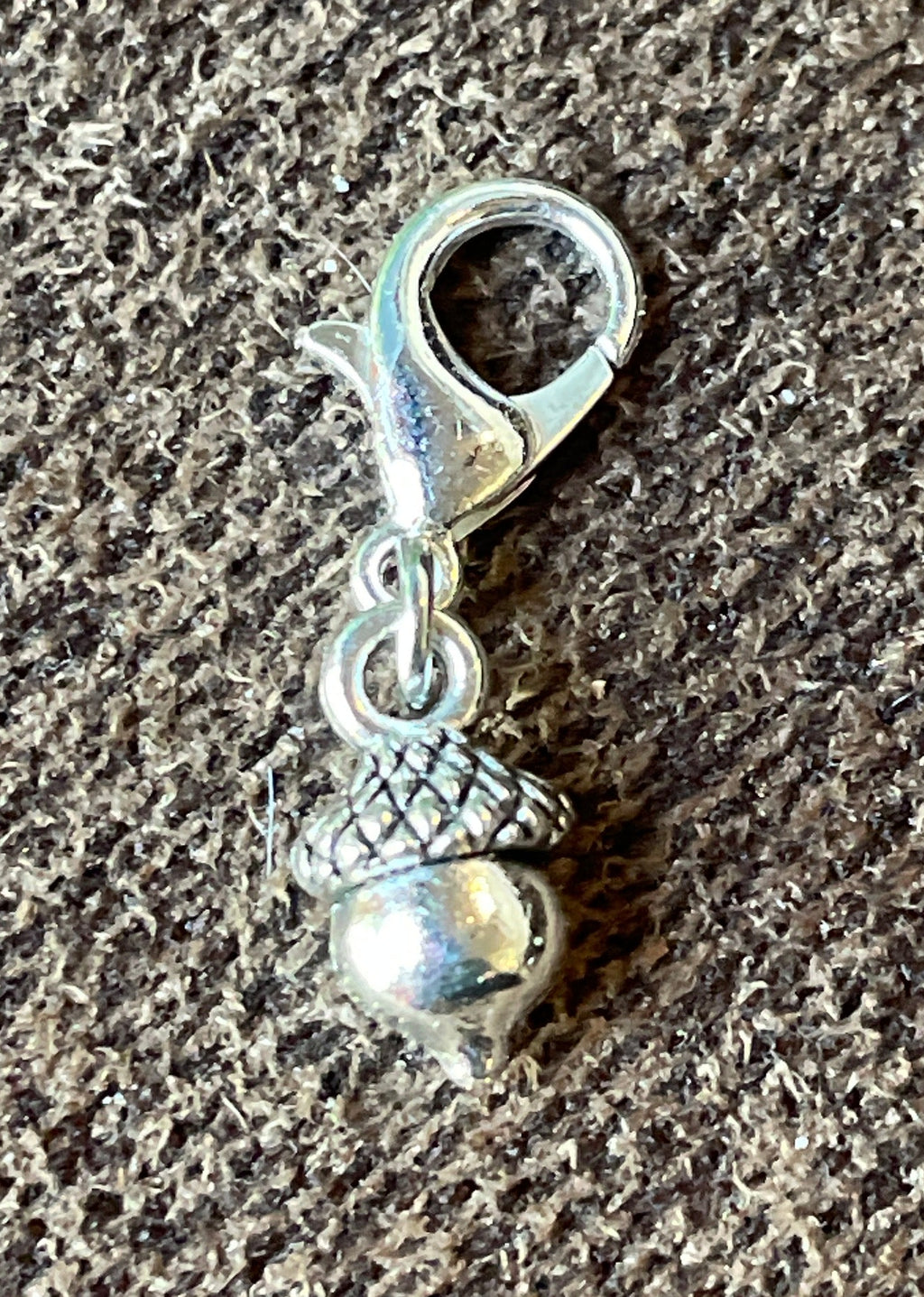A small silver acorn charm with a lobster clasp, resting on a textured gray fabric background.