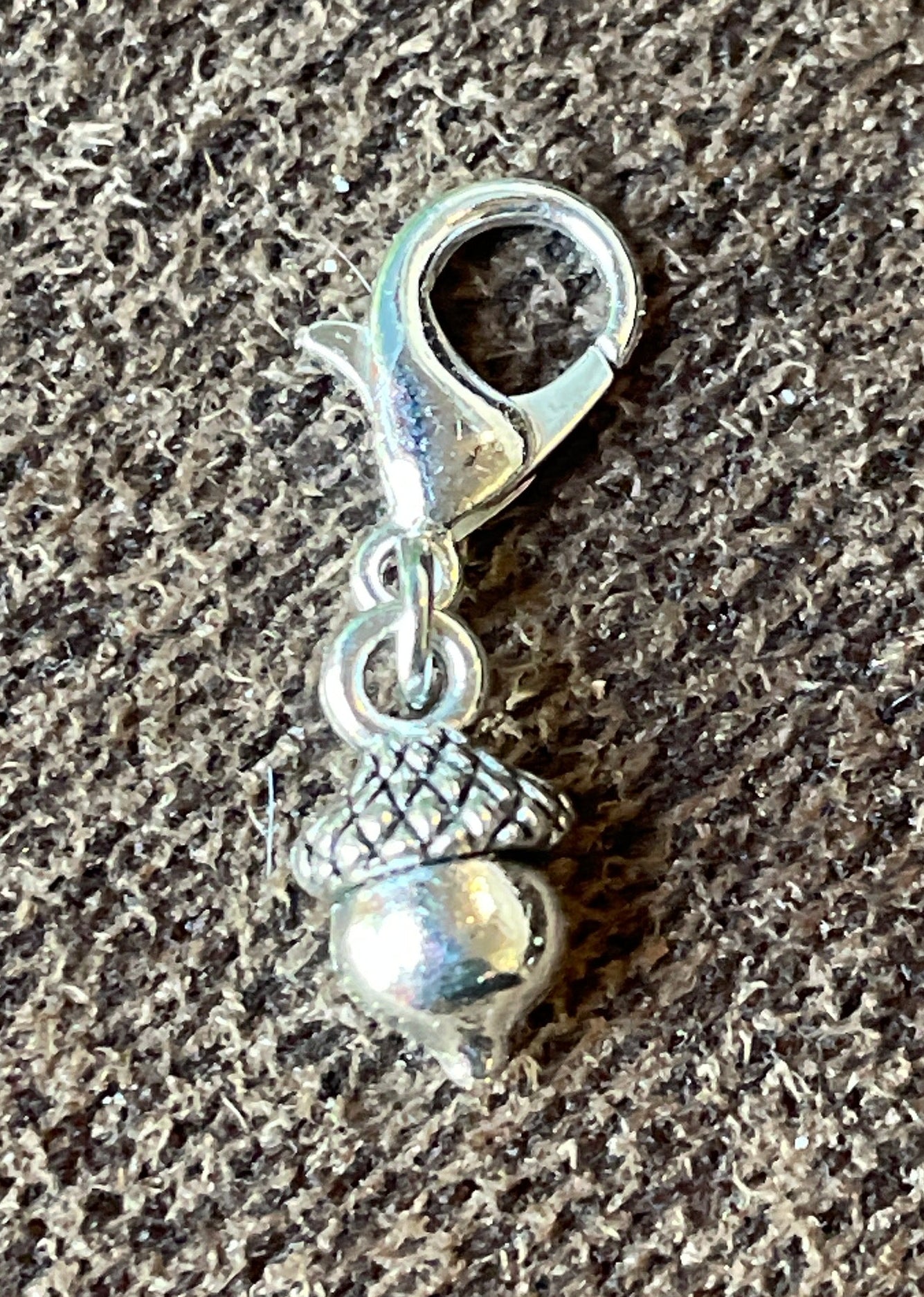 A small silver acorn charm with a lobster clasp, resting on a textured gray fabric background.