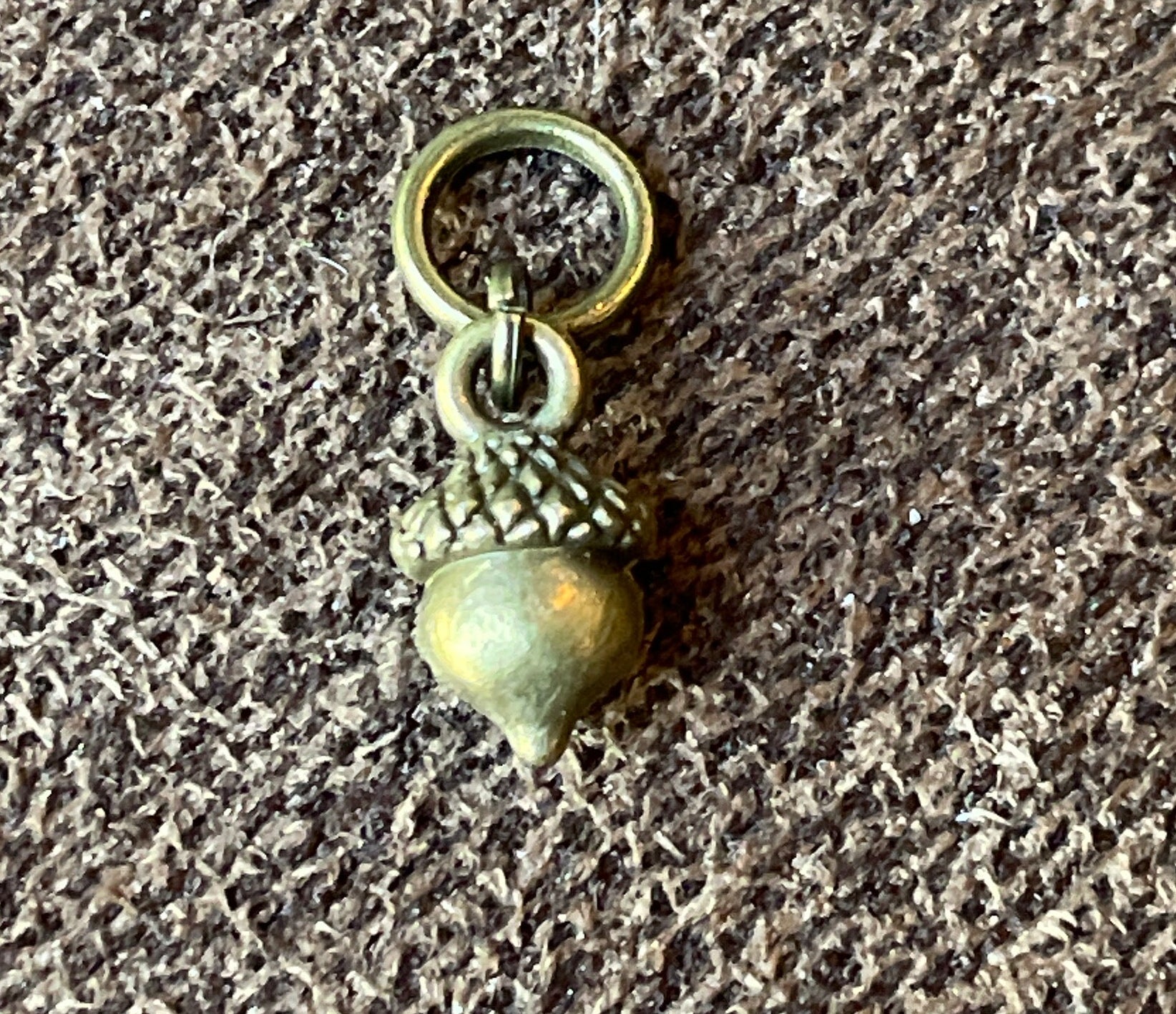 A small metallic acorn charm with a split ring, shown on a textured gray fabric background.