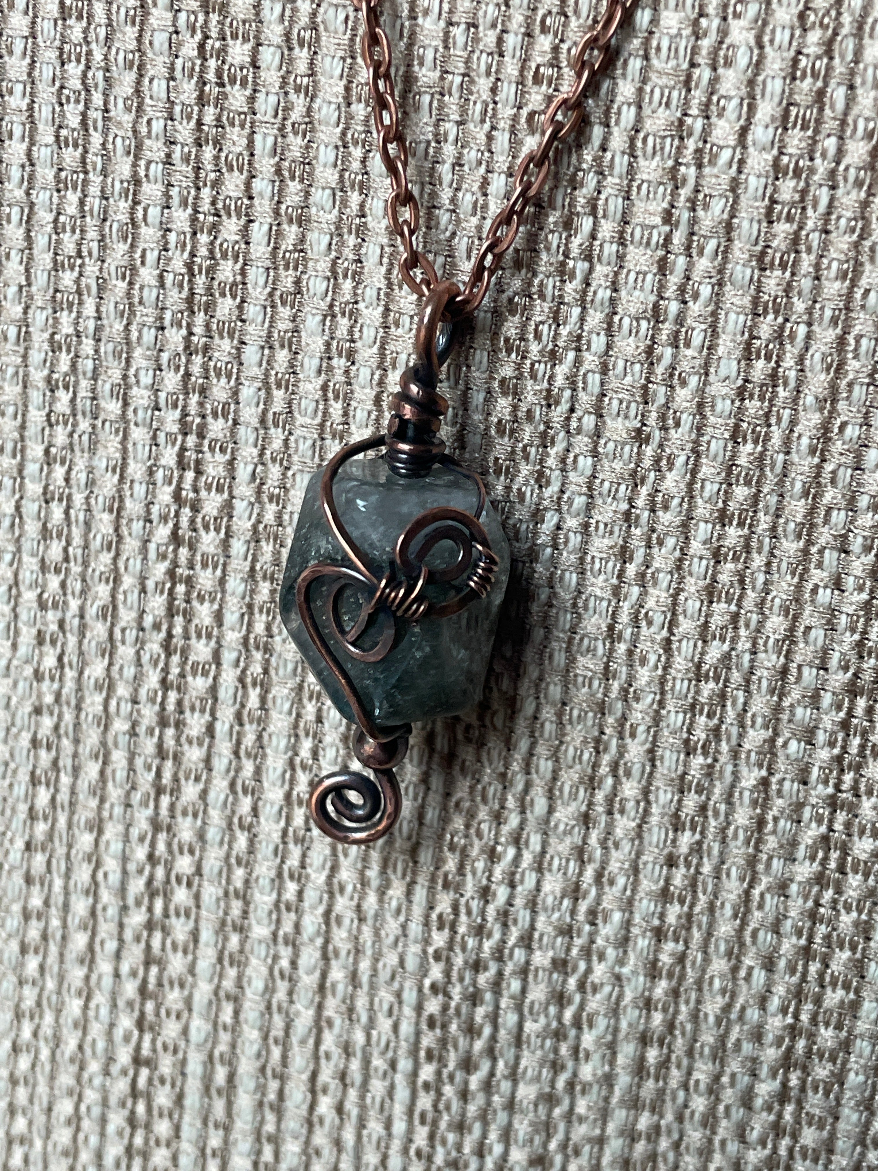 A close-up of a handmade pendant necklace featuring a dark greenish-gray stone wrapped in dark bronze wire, hanging from a matching dark chain on a textured gray background.