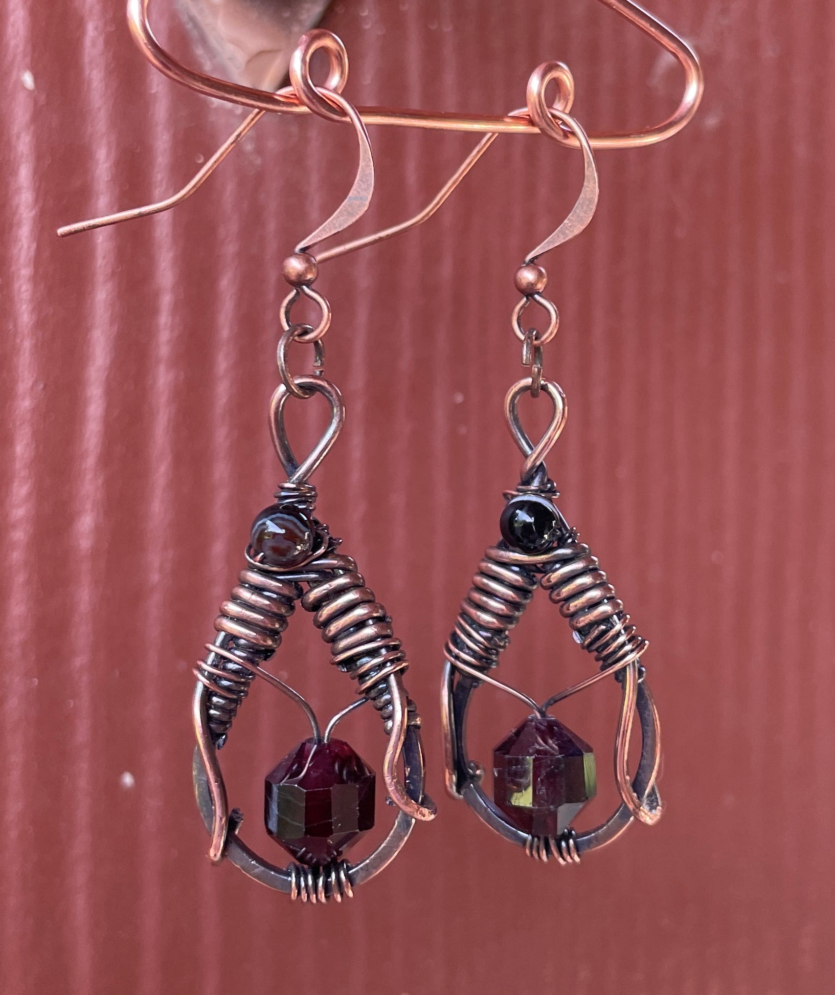 A pair of handmade wire-wrapped earrings with copper-toned hooks, dark metallic wire loops, and faceted deep purple crystal pendants, shown against a textured reddish-brown background.
