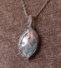 A silver-toned wire-wrapped pendant necklace featuring a natural iridescent blue-green stone, displayed on a textured brown fabric background.