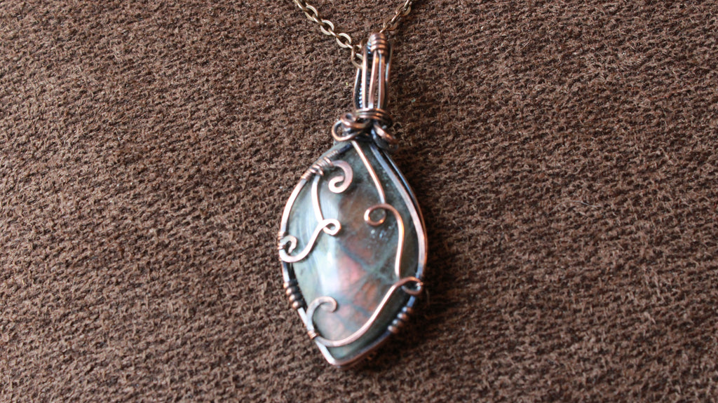 A teardrop-shaped pendant necklace with a silver-toned filigree-wrapped iridescent stone, resting on a textured brown fabric background.
