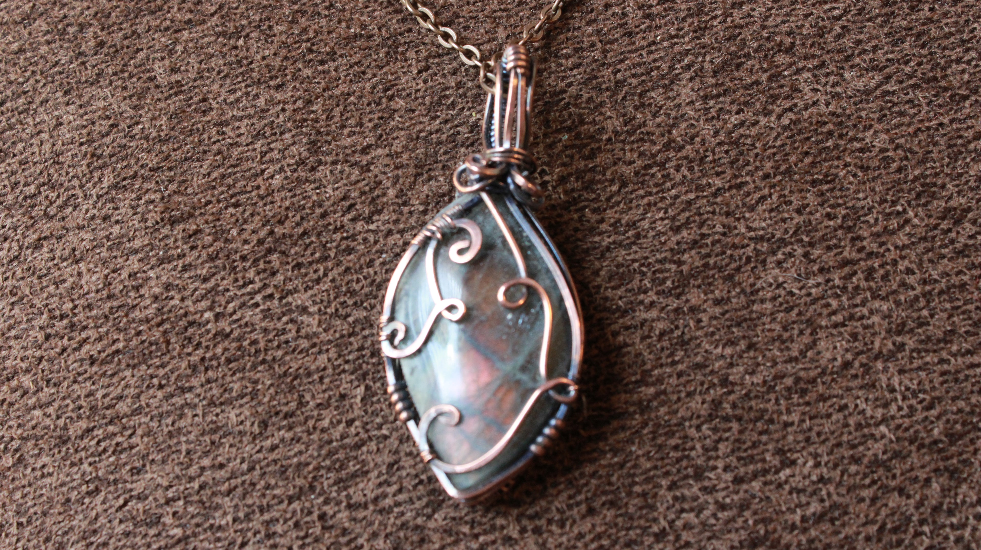A teardrop-shaped pendant necklace with a silver-toned filigree-wrapped iridescent stone, resting on a textured brown fabric background.