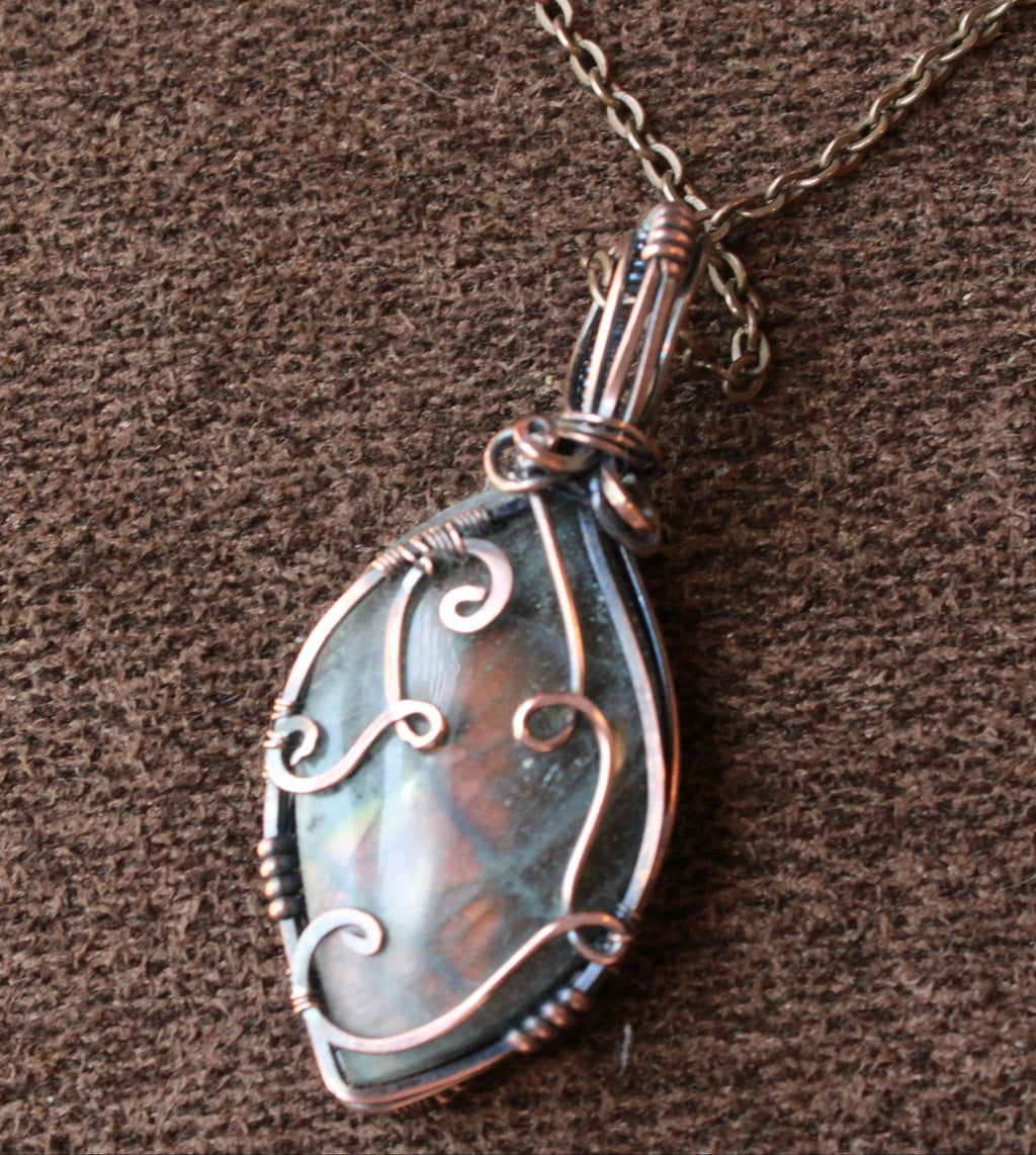 A teardrop-shaped pendant necklace featuring a silver-toned metal chain and a bail wrapped in matching silver wire with decorative scrollwork. The pendant contains an iridescent, pearlescent gray-blue stone.