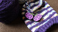 Two silicon stitch stoppers on a set of active knitting needles. The stoppers are  purple  and black pumpkins with the flying bats across them.