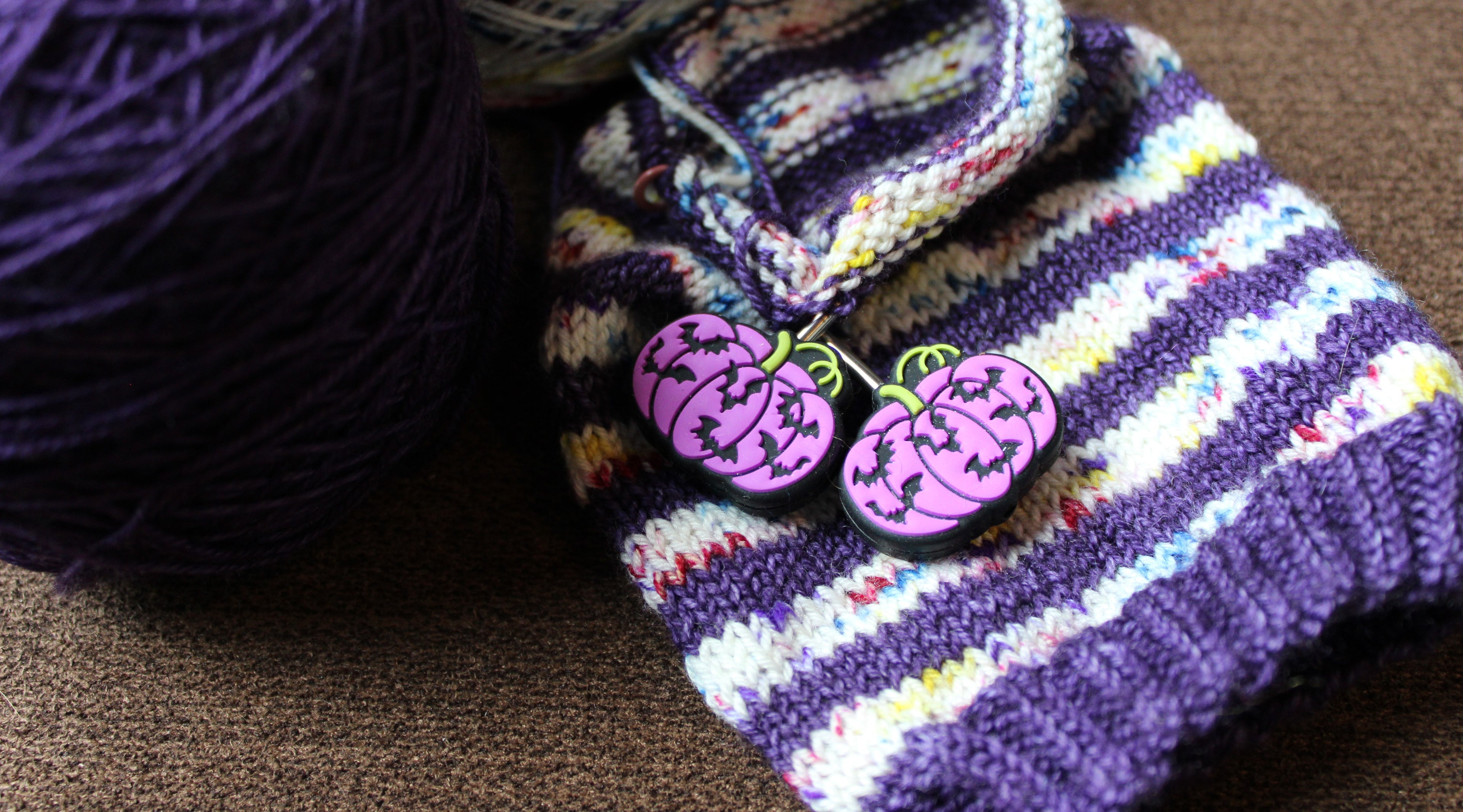 Two silicon stitch stoppers on a set of active knitting needles. The stoppers are  purple  and black pumpkins with the flying bats across them.