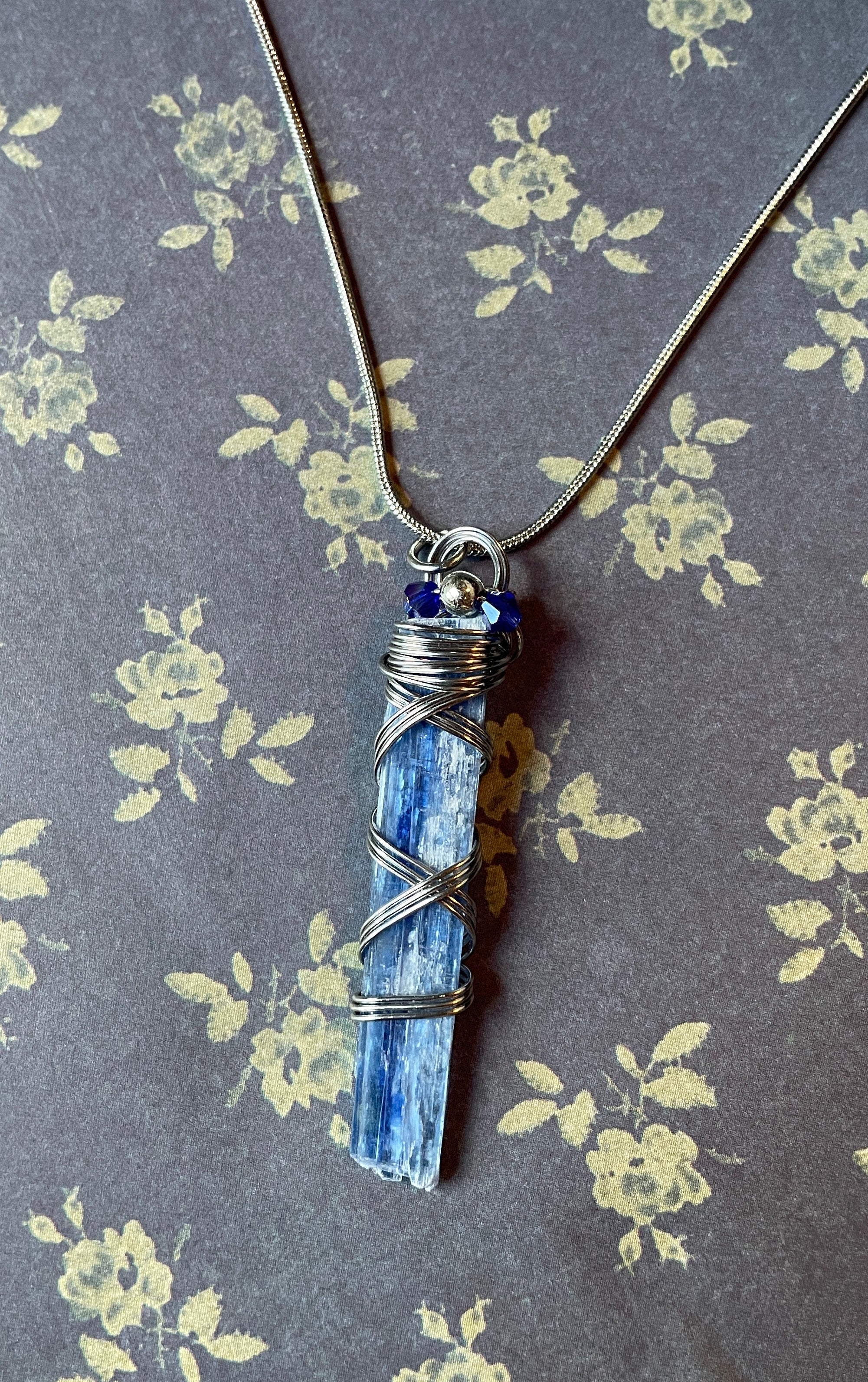A handmade silver wire-wrapped necklace featuring a blue sapphire point pendant, displayed on a dark gray floral-patterned background.