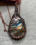 A close-up of a hand holding a teardrop-shaped wire-wrapped pendant necklace with a shimmering iridescent stone. The pendant features copper-toned wire wrapping and a matching chain, set against a textured gray background.