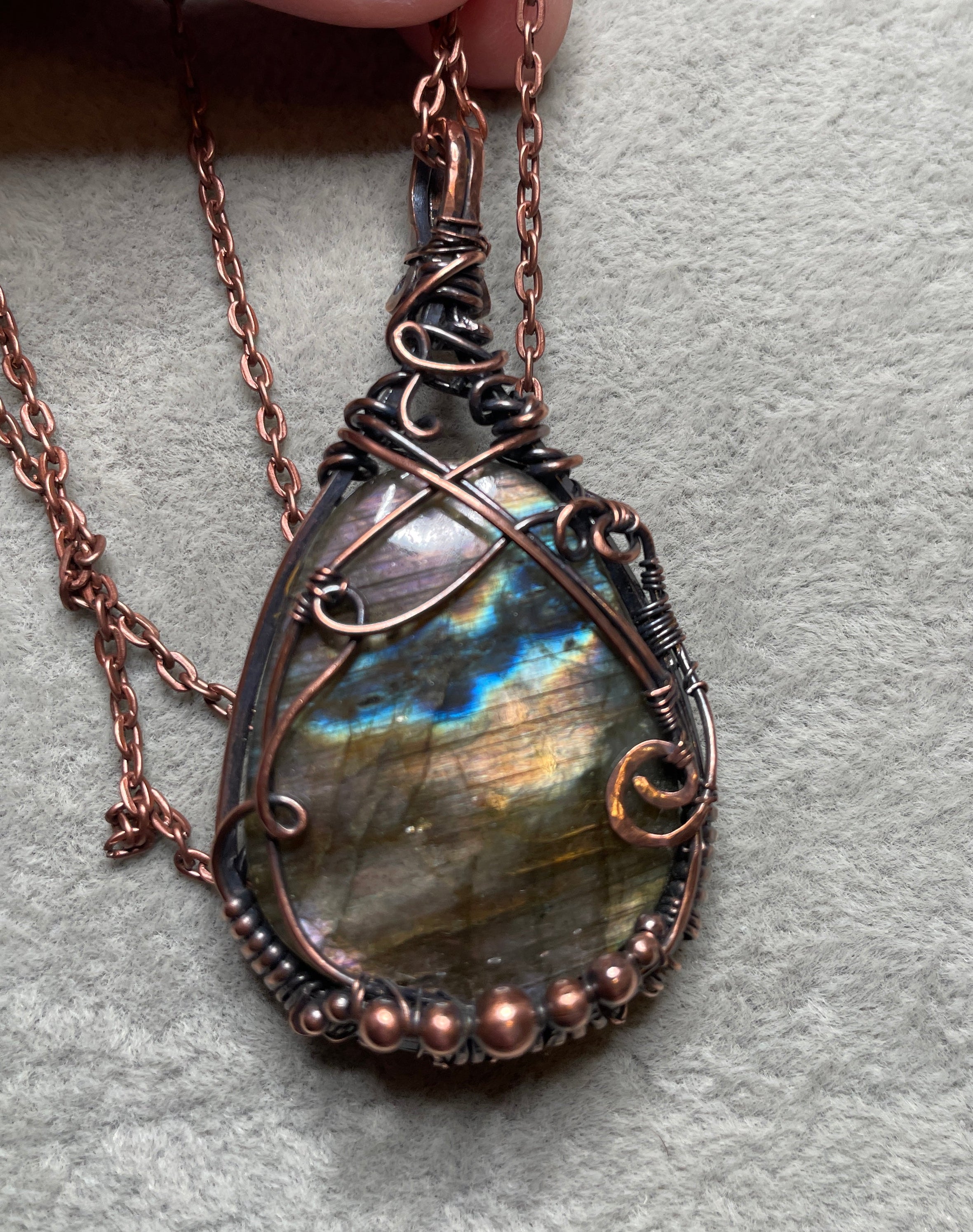 A close-up of a hand holding a teardrop-shaped wire-wrapped pendant necklace with a shimmering iridescent stone. The pendant features copper-toned wire wrapping and a matching chain, set against a textured gray background.