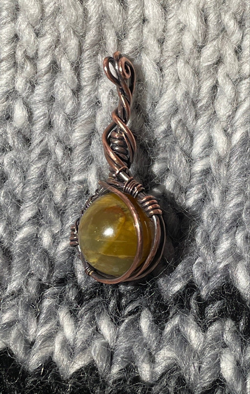 A small handmade pendant with a copper wire bail and a round olive-green stone, resting on a gray and black textured knit blanket.