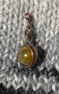 A small handmade pendant with a copper wire bail and a round olive-green stone, resting on a gray and black textured knit blanket.
