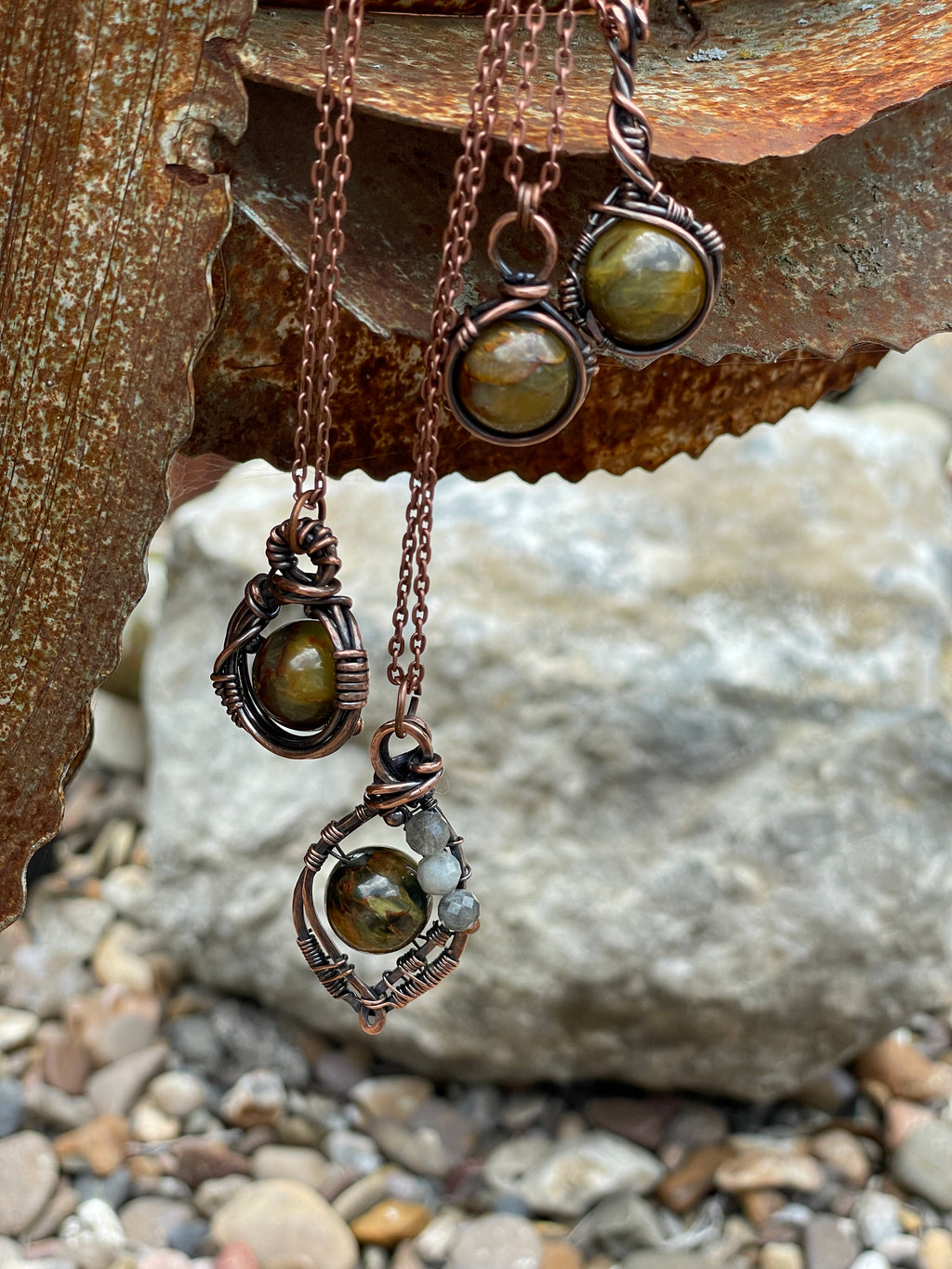 Copper wire-wrapped jewelry set featuring three pendant necklaces and two matching drop earrings with round greenish-brown stones, displayed against a rustic rusty metal sheet and stone background.