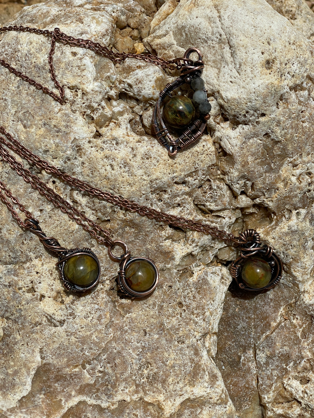 Four handmade wire-wrapped pendant necklaces with round olive-green gemstone pendants and dark brown chains, displayed on a textured beige rock surface.