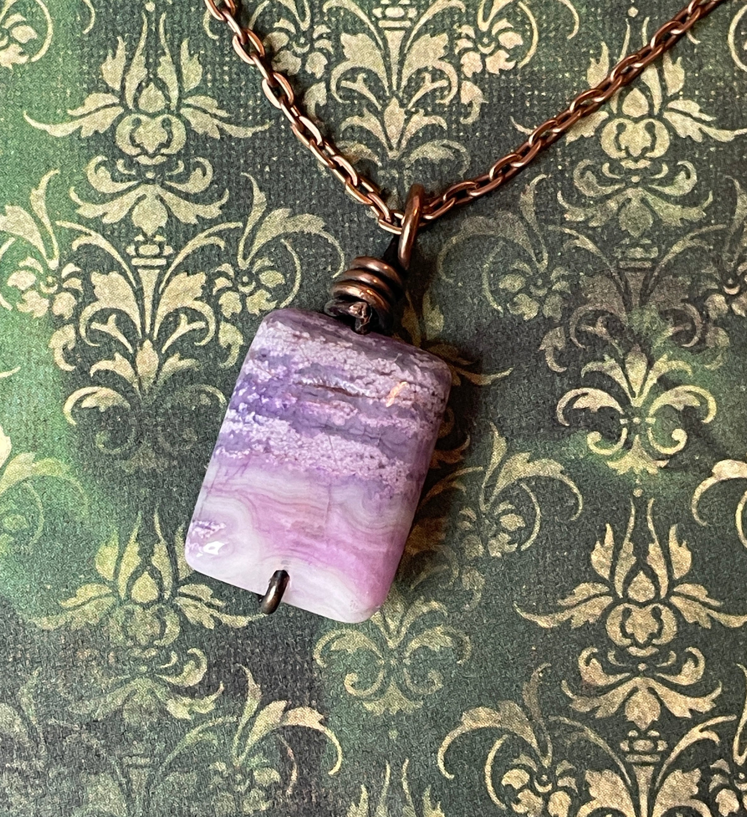 A handmade pendant necklace featuring a rectangular purple and lavender stone with white marbling, wrapped in copper wire, hanging from a copper chain against a green damask patterned background.