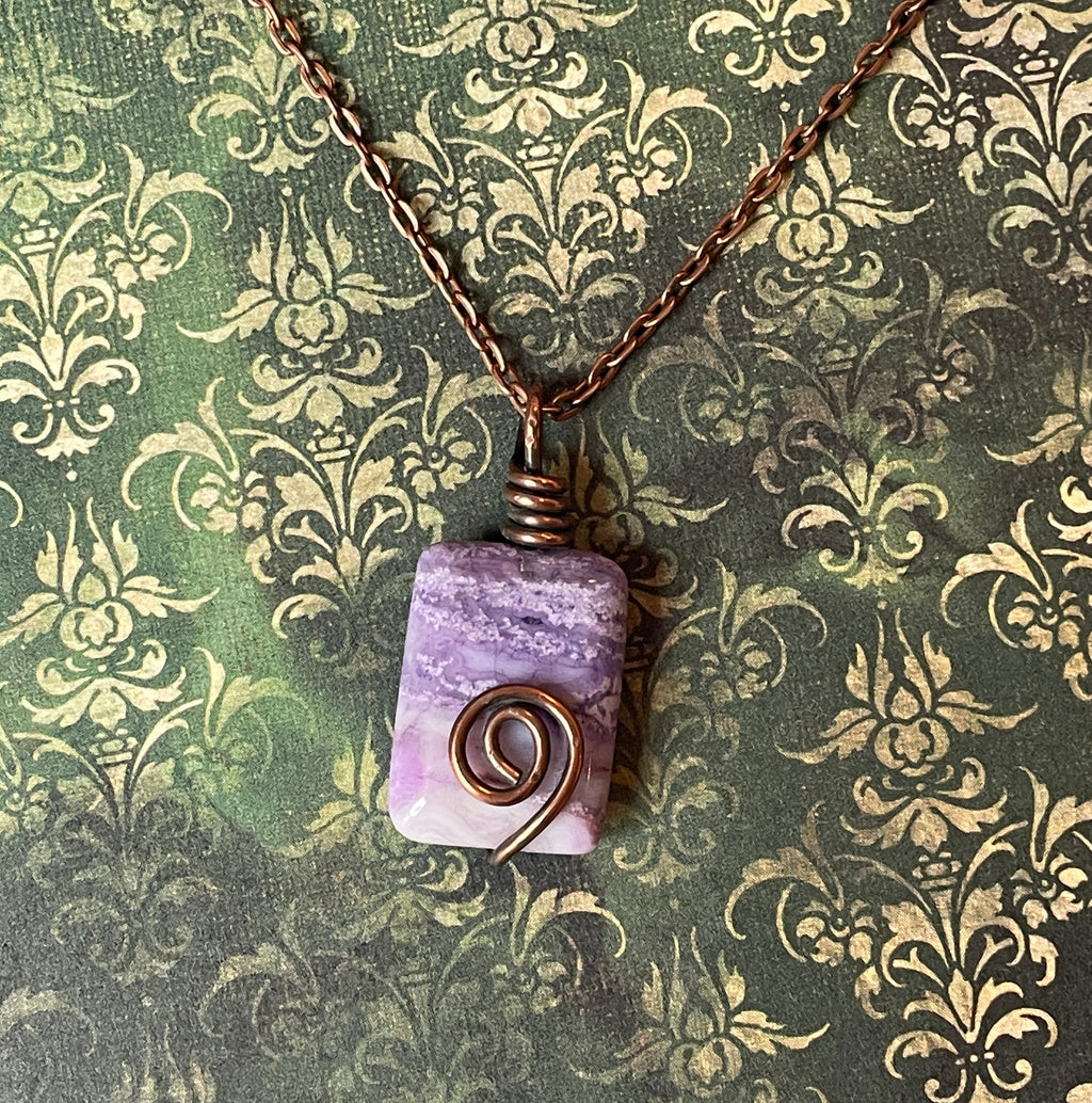 A handmade wire-wrapped necklace featuring a rectangular lavender and purple stone pendant with a copper spiral accent, hanging from a copper chain against a green damask patterned background.