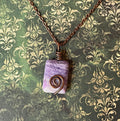 A handmade wire-wrapped necklace featuring a rectangular lavender and purple stone pendant with a copper spiral accent, hanging from a copper chain against a green damask patterned background.