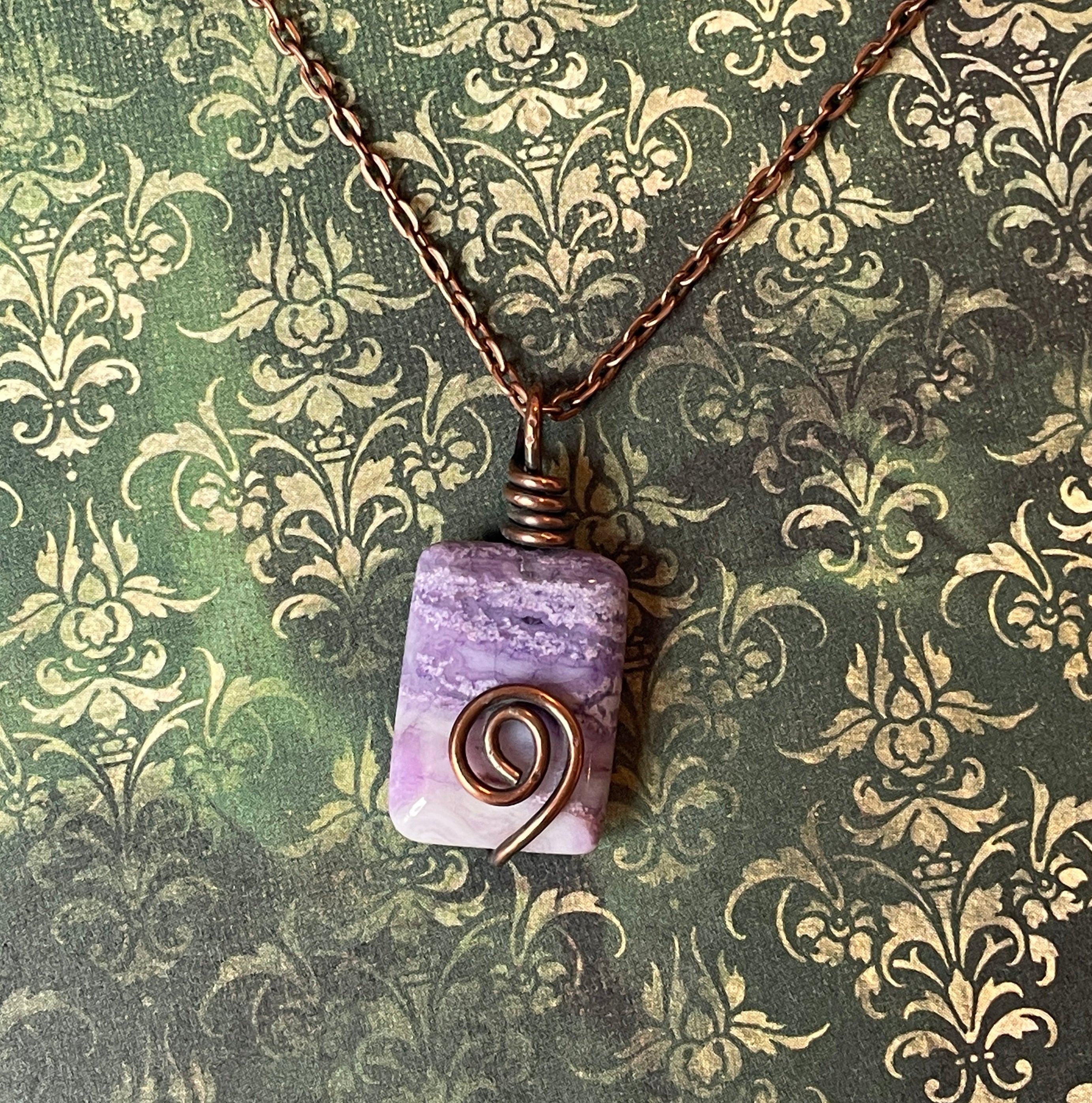 A handmade wire-wrapped necklace featuring a rectangular lavender and purple stone pendant with a copper spiral accent, hanging from a copper chain against a green damask patterned background.