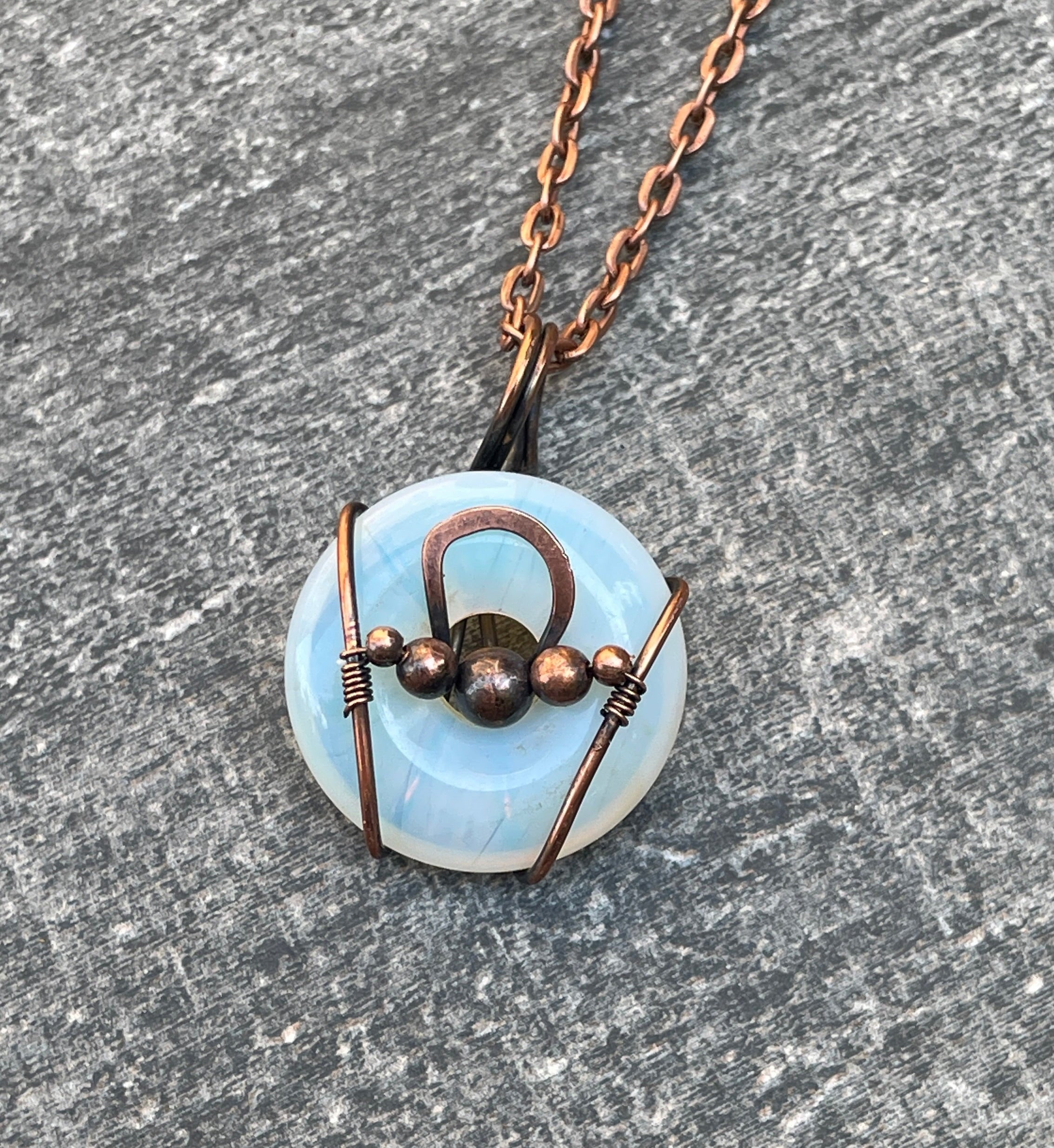 A handmade pendant necklace featuring a light blue circular stone wrapped in copper wire with a central copper loop and bead details, resting on a textured gray stone surface.